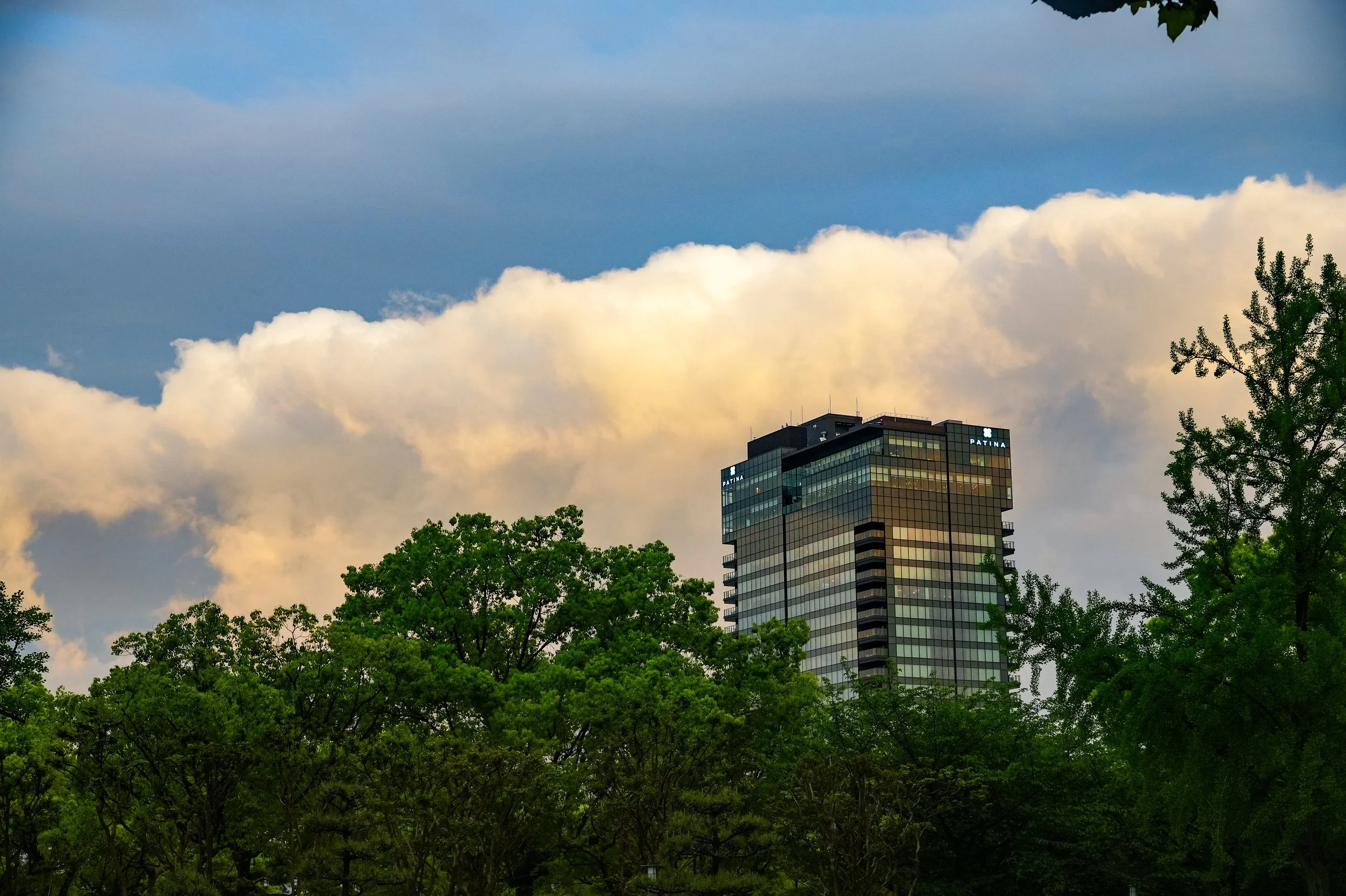 A high-rise building with a blue and black exterior, buildings logo on top right corner, surrounded by green trees, under a cloudy sky.
