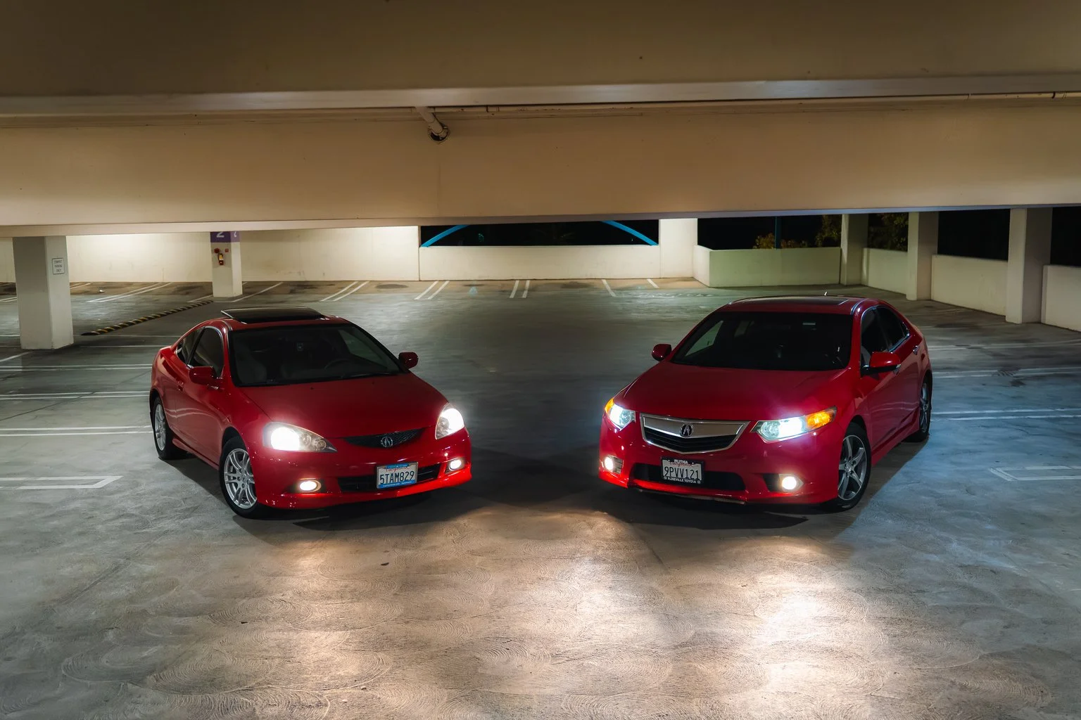 Two red cars parked side by side in a dimly lit underground parking garage, one with its headlights on.