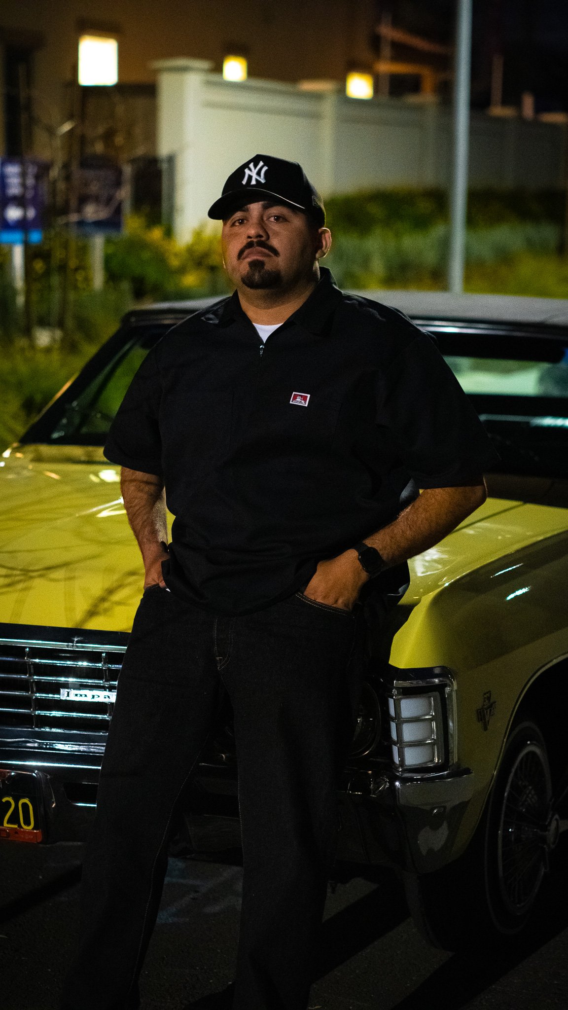 A man standing next to a yellow vintage car at night, wearing a black shirt and a black New York Yankees cap.