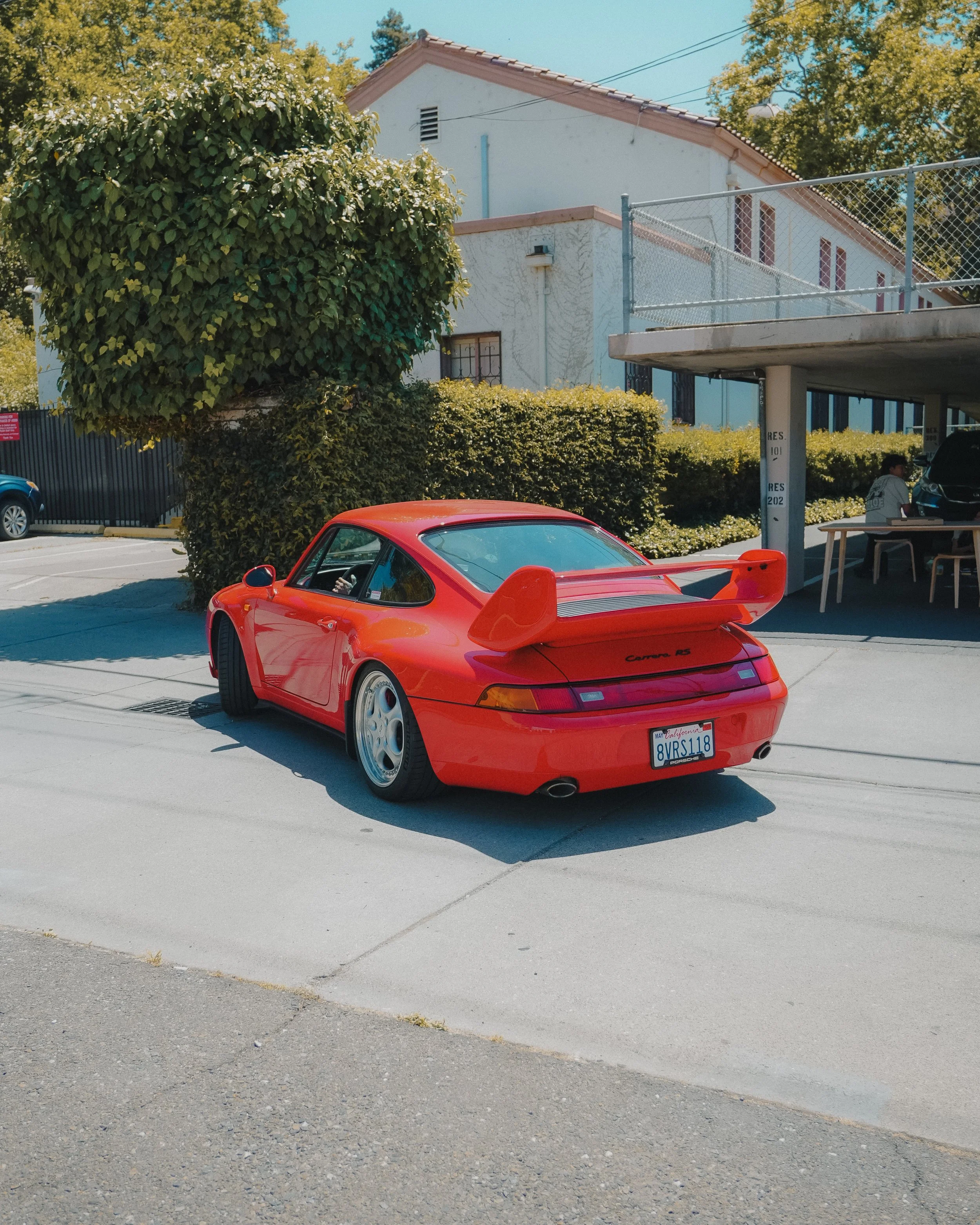 A red Porsche Carrera RS parked in an outdoor parking lot near a white building with red-tiled roof. There is a person sitting on a bench under a parking shelter, and a large green leafy tree next to the building.