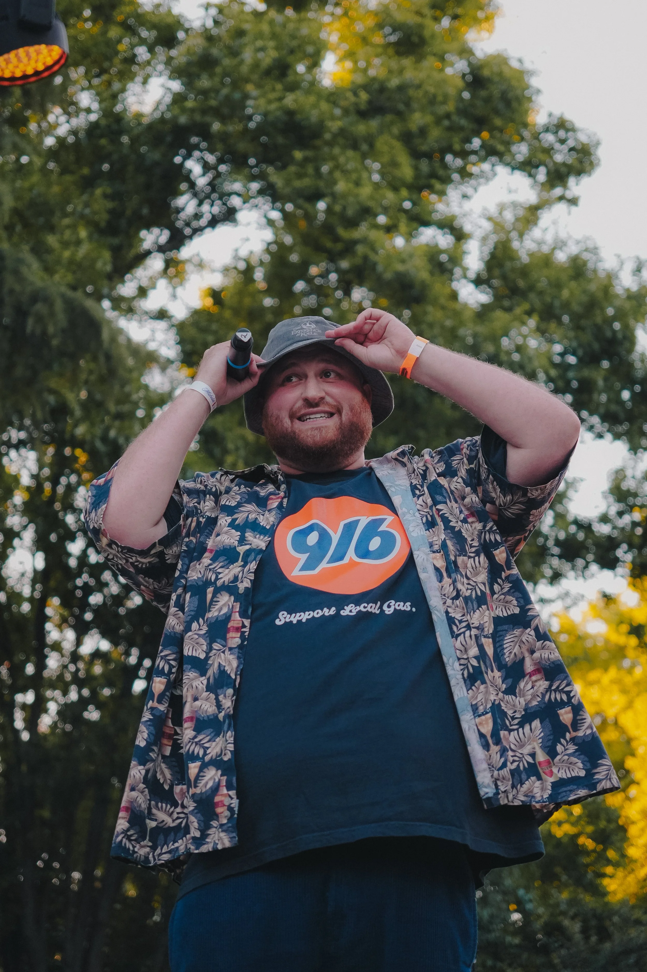 A smiling man with a beard wearing a gray hat, floral shirt, and a dark T-shirt with a 9/16 logo and support local gas text, standing outdoors in front of trees with sunlight filtering through the leaves.