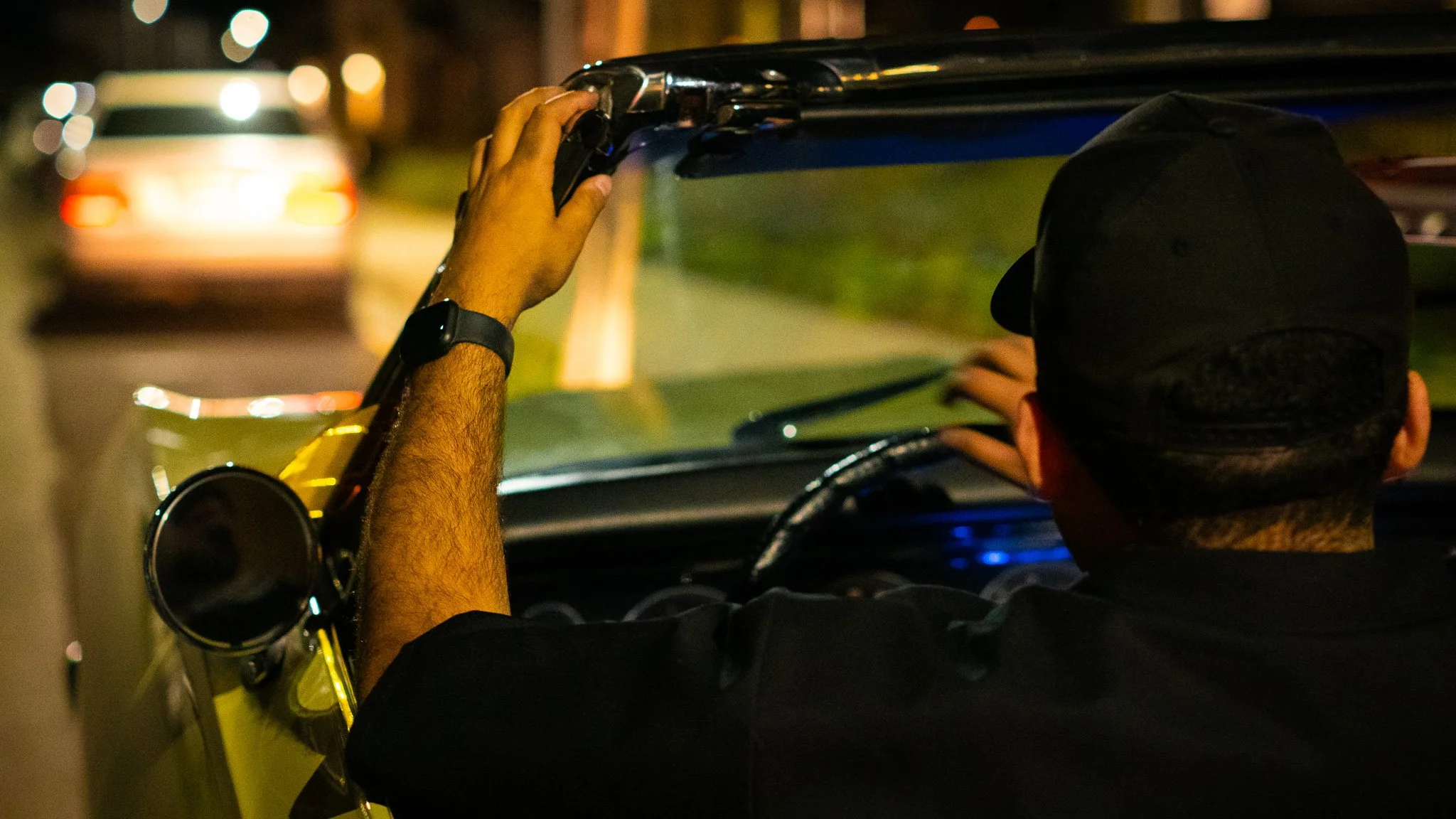 A person wearing a black cap and black shirt is seen from behind, holding a smartphone to the dashboard of a car, at night. The car's interior is dark, and the background shows blurred lights from other vehicles.