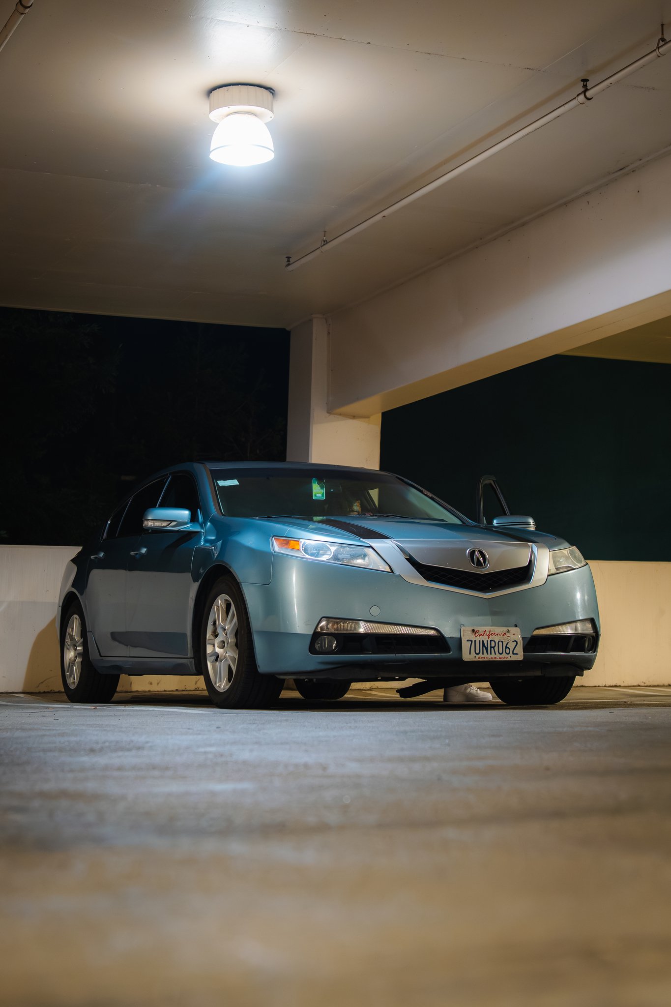 A blue Acura sedan parked in an underground parking garage at night, with surrounding concrete walls and a bright ceiling light.