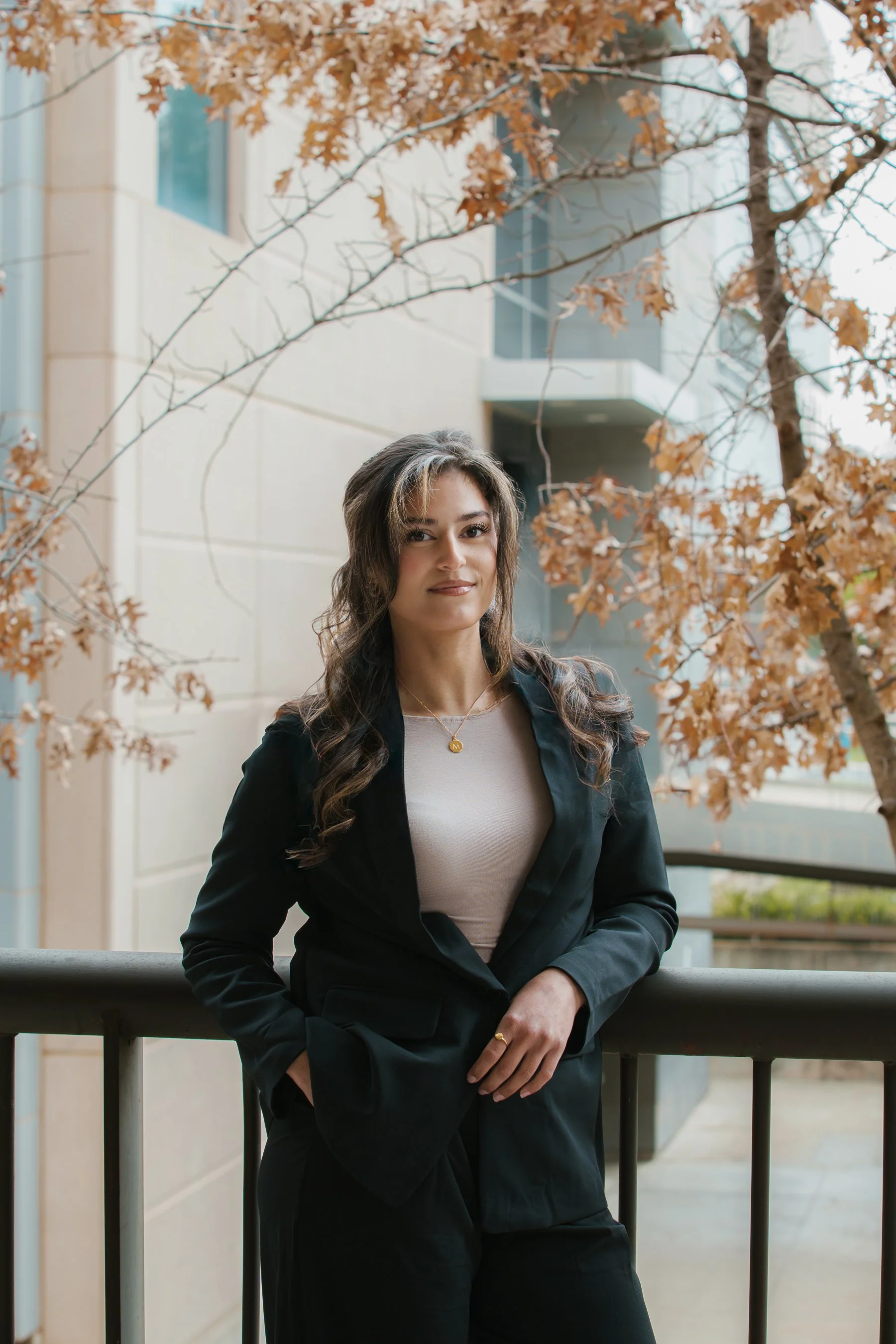 A woman with long, wavy brown hair, wearing a black blazer and a beige top, standing outdoors on a balcony during fall. She is leaning against a railing with one hand in her pocket, with a background of a building and a tree with brown autumn leaves.
