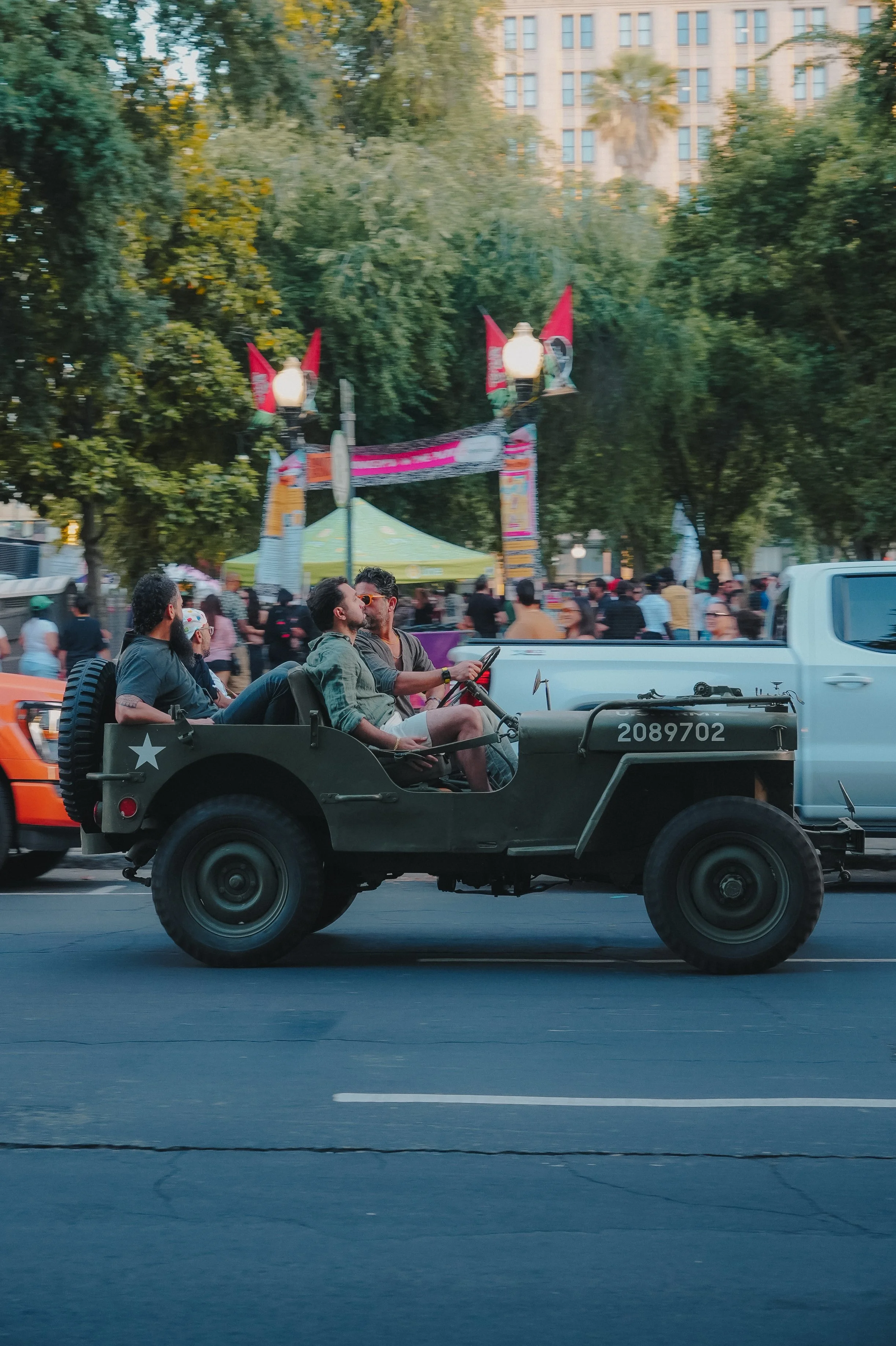 A street parade scene with two men sitting in a military-style vehicle driving on a city street. The vehicle has a star emblem and the number 2089702 on it. In the background, there is a crowd of people, trees, and a partly visible multi-story buildi