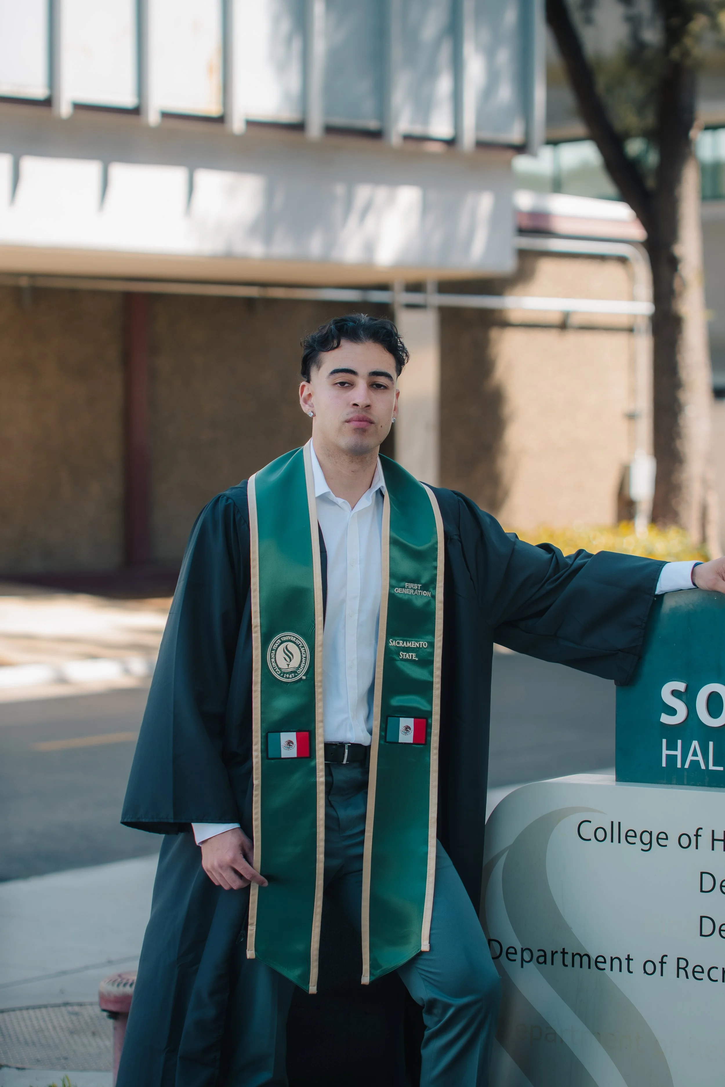 A young man in a graduation gown holding a cap standing outdoors near a sign for the College of Humanities.