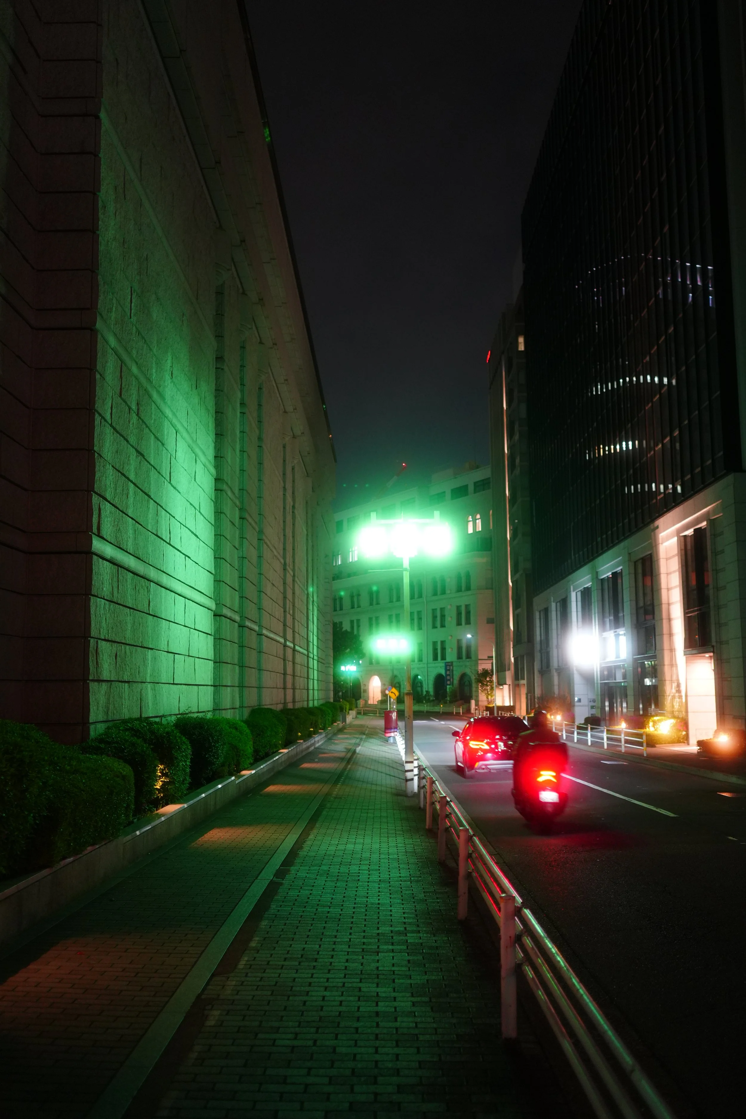 Nighttime city street scene with a sidewalk, green lighting on a stone building, and traffic with red tail lights, tall buildings on the right, and a dark sky.