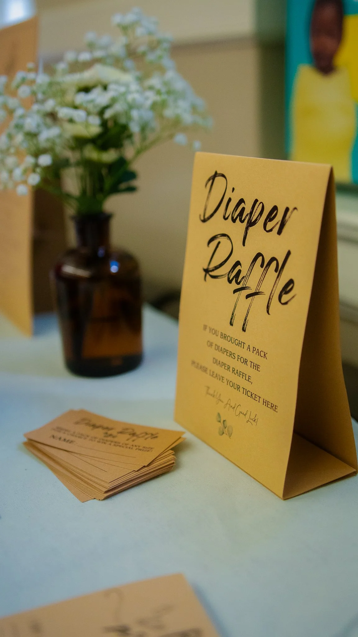 A yellow sign promoting a diaper raffle with text instructing to bring a pack of diapers for a raffle and leave the ticket, with a blurred background showing a brown vase with white flowers and a colorful piece of artwork.