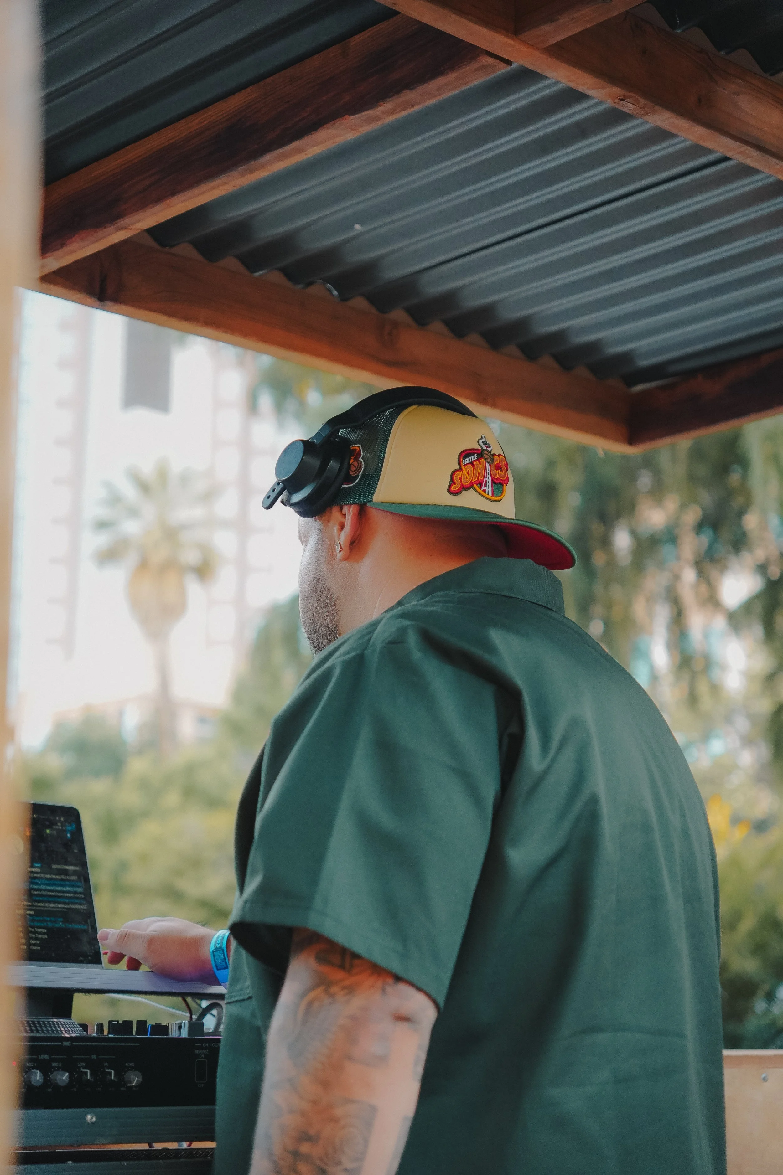 A person wearing a San Francisco 49ers cap with headphones, working at a DJ or music setup with a laptop and audio equipment, outdoors with trees and buildings in the background.