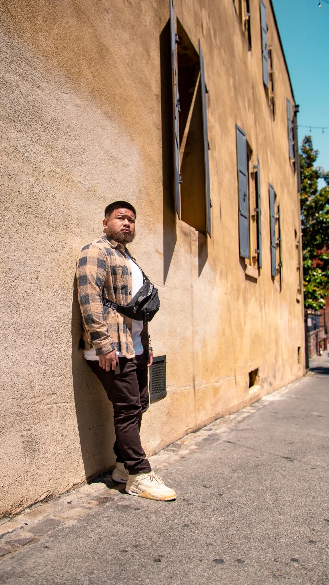 A man with a beard and short hair leaning against a beige stucco wall with closed black window shutters on a sunny day. He is wearing a brown plaid jacket, white t-shirt, dark pants, and cream-colored sneakers. The street has cobblestone and pavement