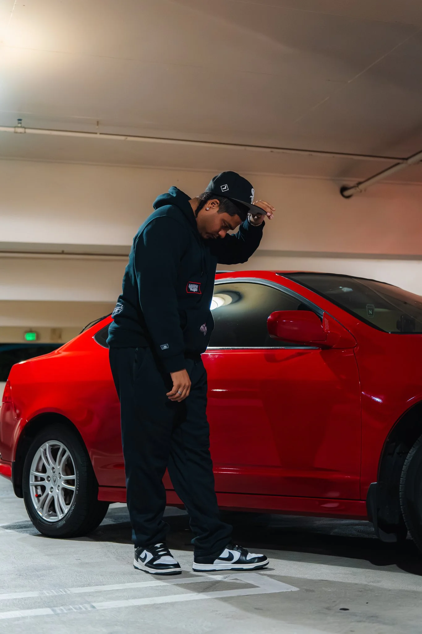 A young man in a black hoodie and black Nike sneakers standing next to a red car in an underground parking garage, looking down with a hat on.