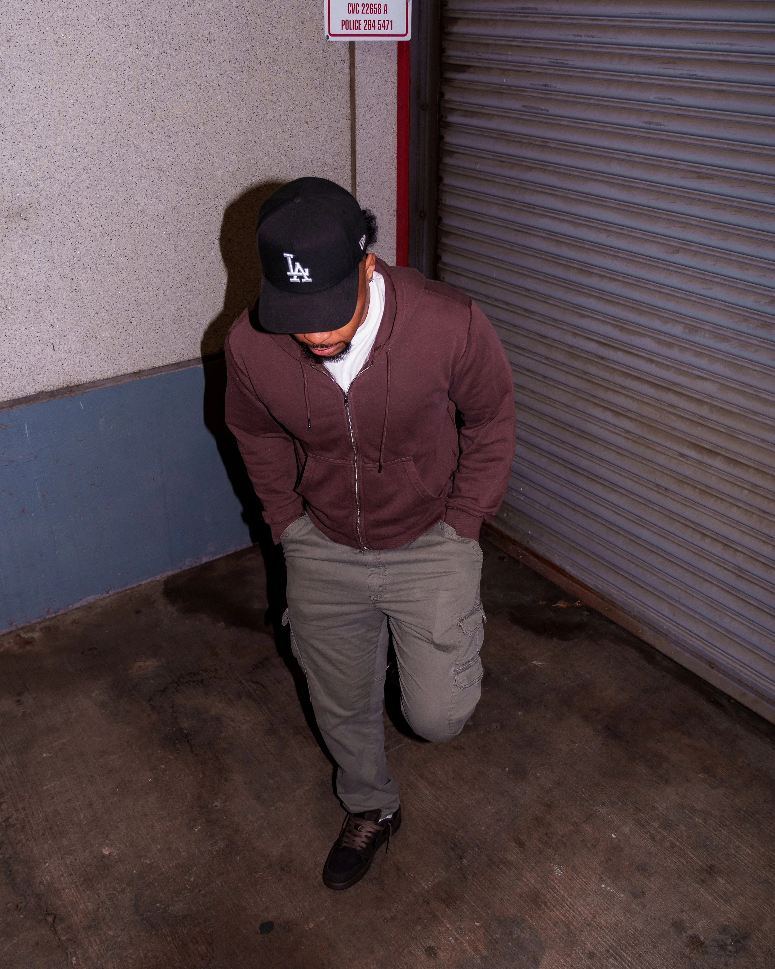 A young man wearing a black LA Dodgers baseball cap, brown zip-up hoodie, gray cargo pants, and black sneakers with orange soles, standing against a concrete wall and a metal roller shutter door.