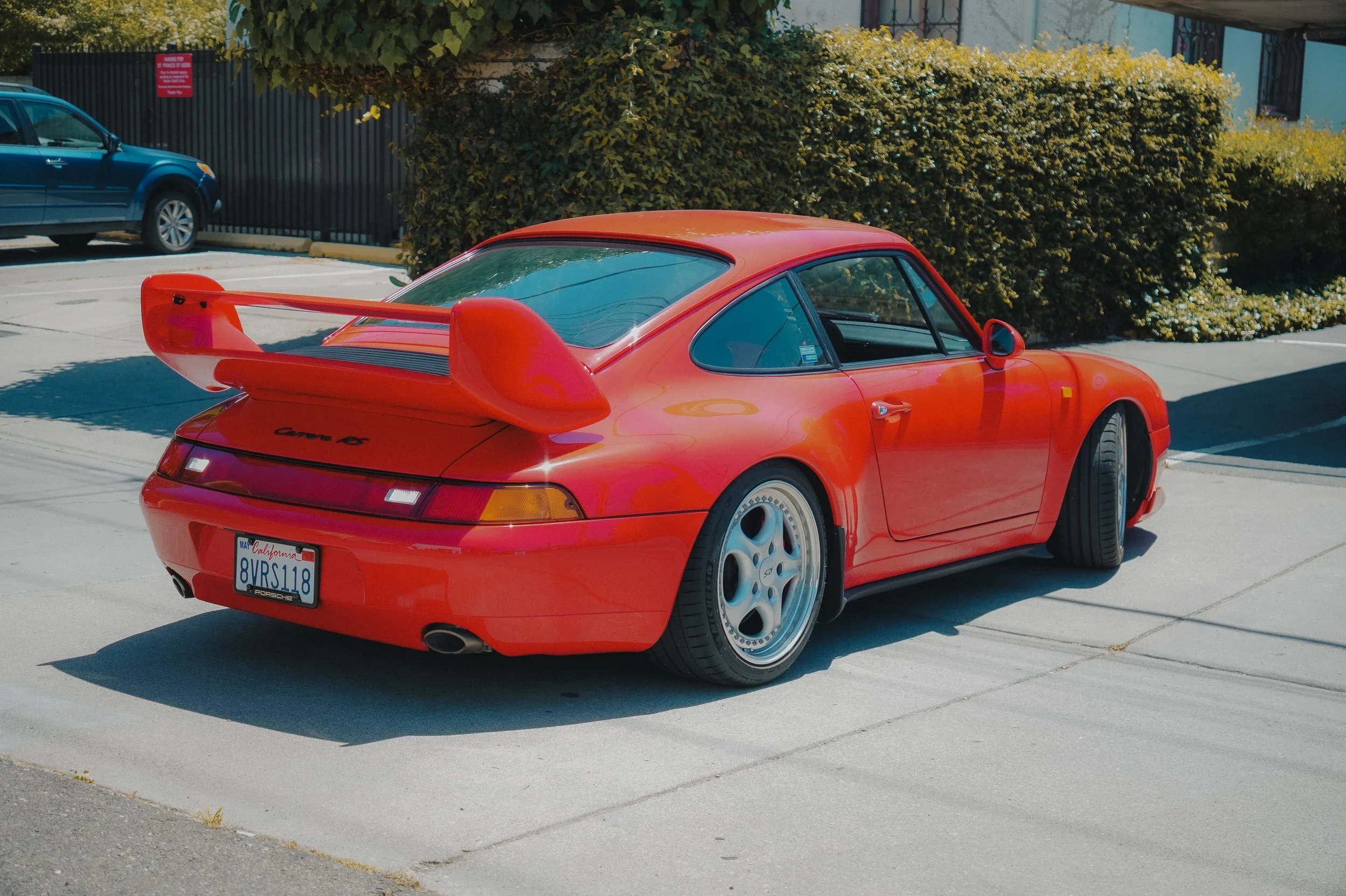 Red Porsche 911 sports car parked in a parking lot with another vehicle and bushes in the background.