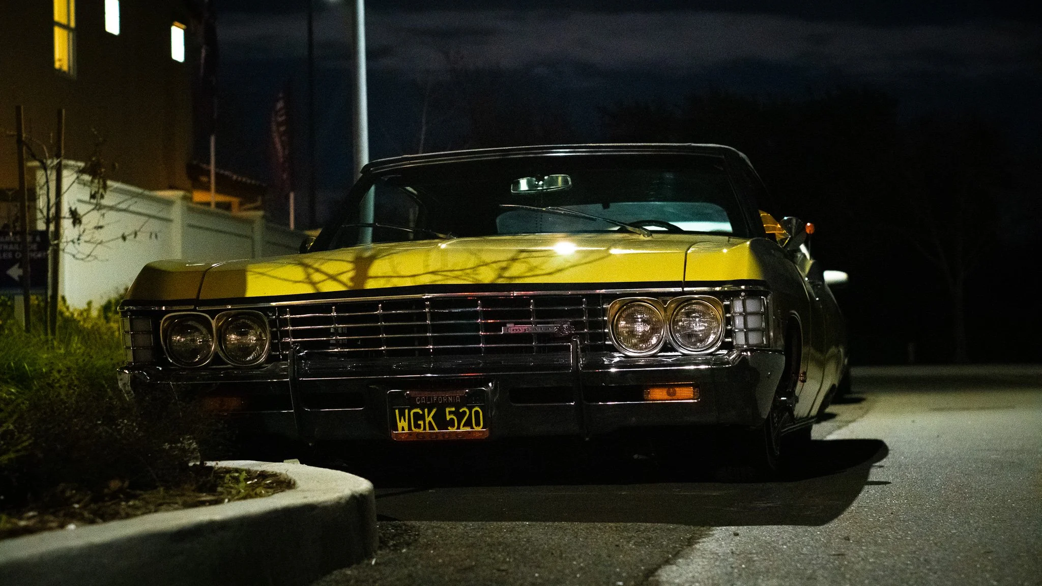 A vintage black and yellow car parked on the side of a street at night, illuminated by surrounding lights, with a white fence, leafless trees, and a building in the background.