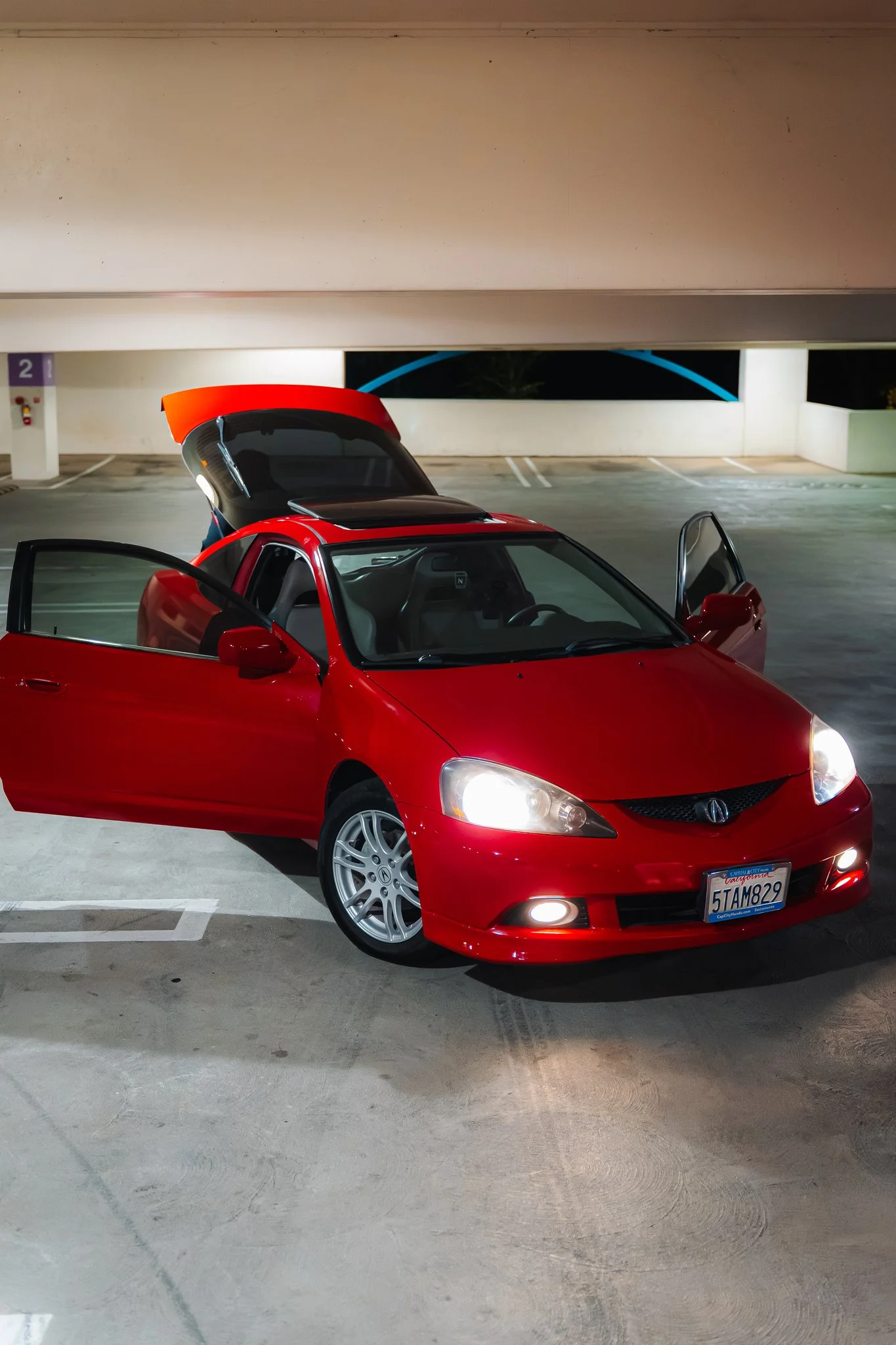 A red sports car parked in a mostly empty parking garage at night, with open doors and the trunk raised. The car is a Honda with a California license plate, under artificial lighting.