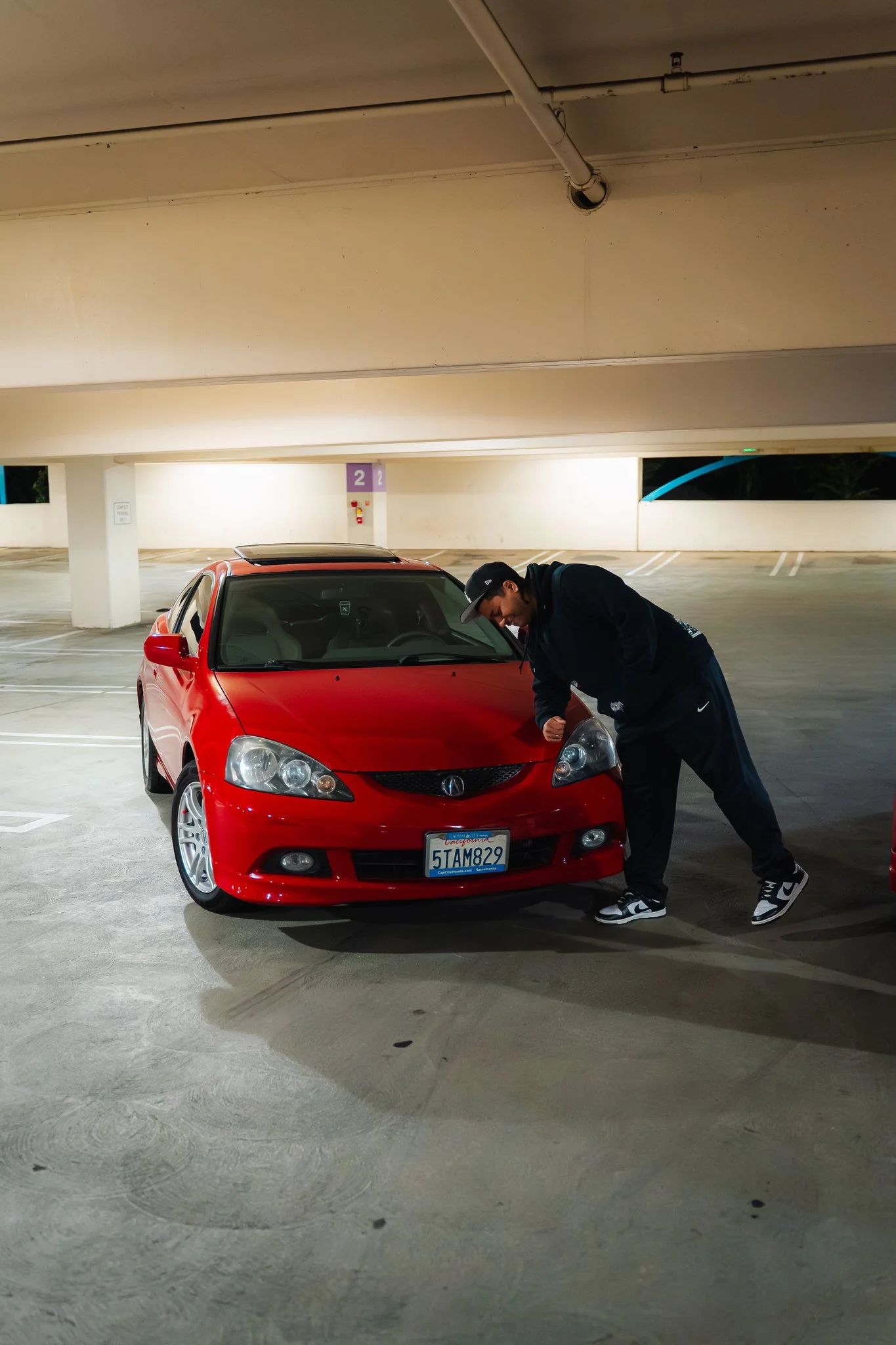 A man in a black hoodie, black pants, and a black cap leans over a red sports car with a California license plate, parked in an empty parking garage at night.