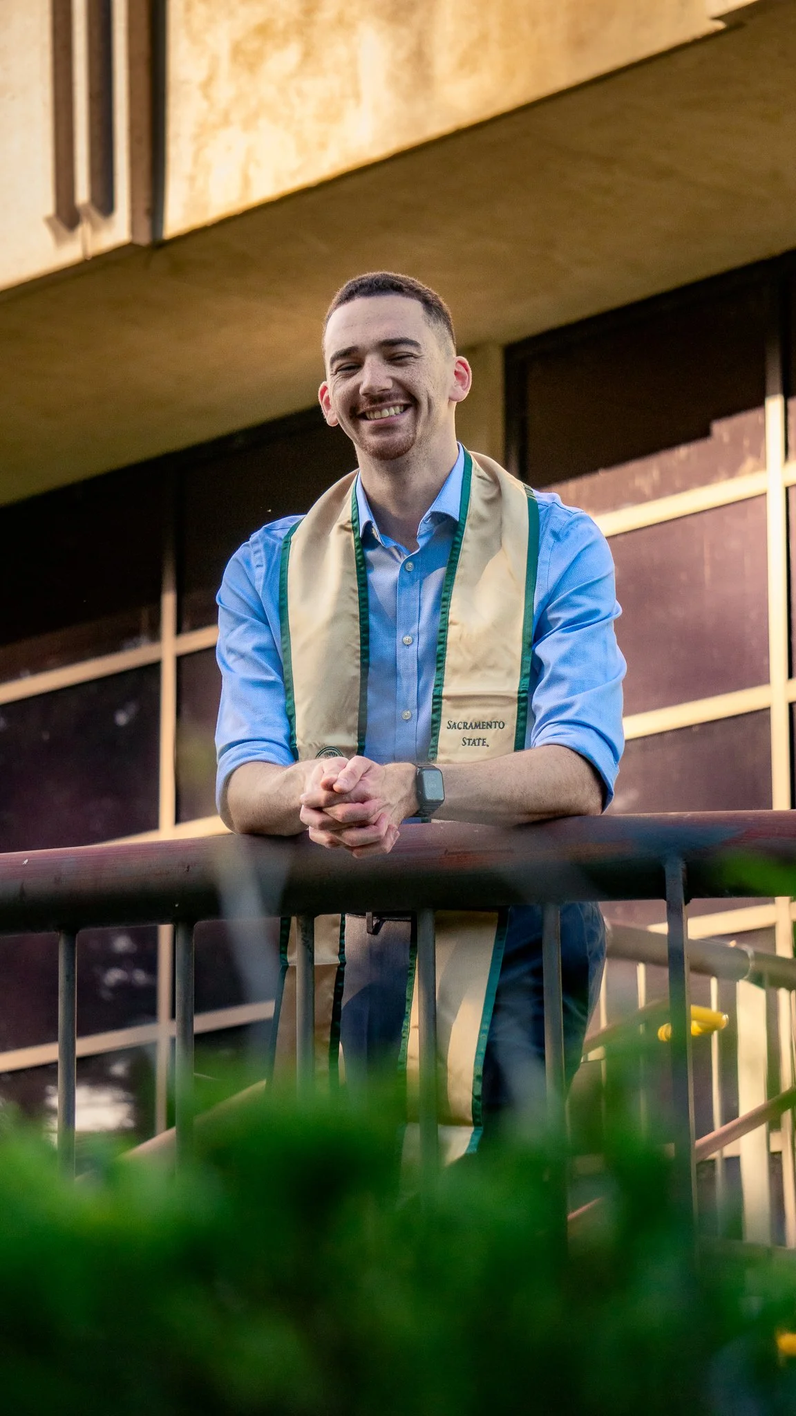 A young man smiling, wearing a blue shirt and a light-colored sash with 'Sacramento State' written on it, standing behind a railing in an indoor or outdoor setting.