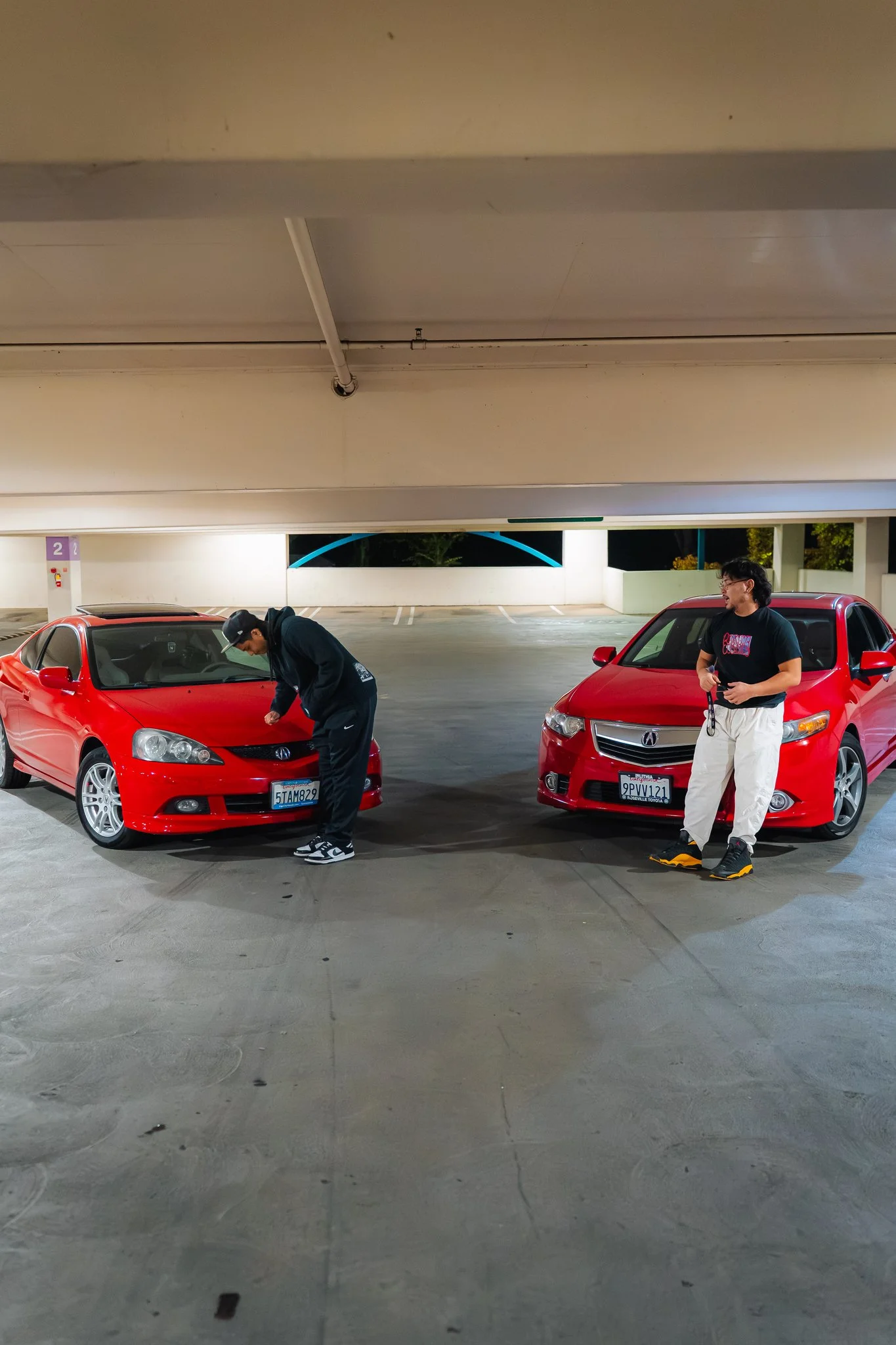 Two young men with black hair stand near two red cars in a parking garage. The parking garage has a concrete floor and ceiling with purple parking space signs. One man is wearing a black hoodie, black pants, and sneakers, looking into the front of on
