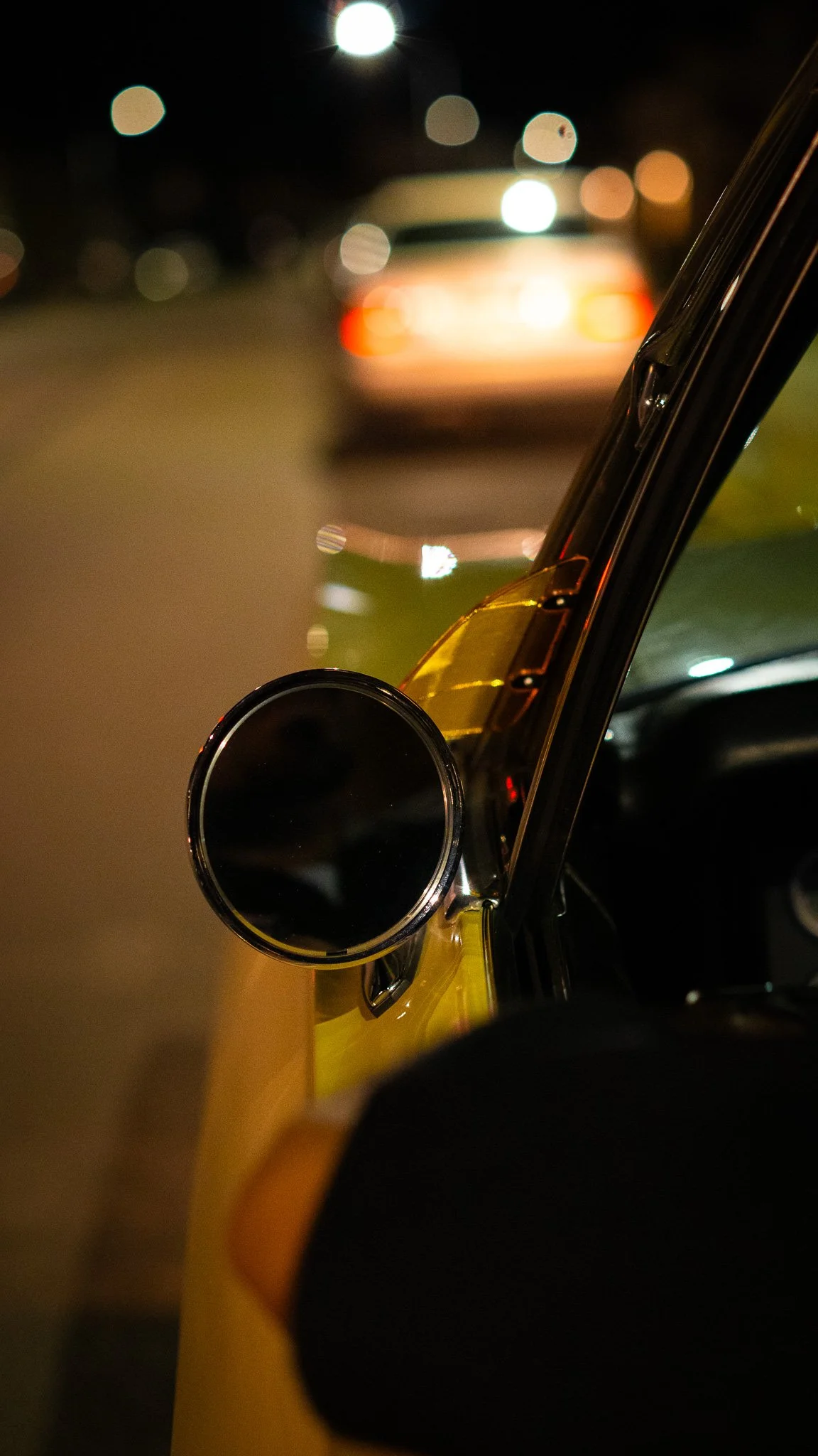 Close-up of a yellow car's side mirror at night, with blurred lights and a vehicle in the background.