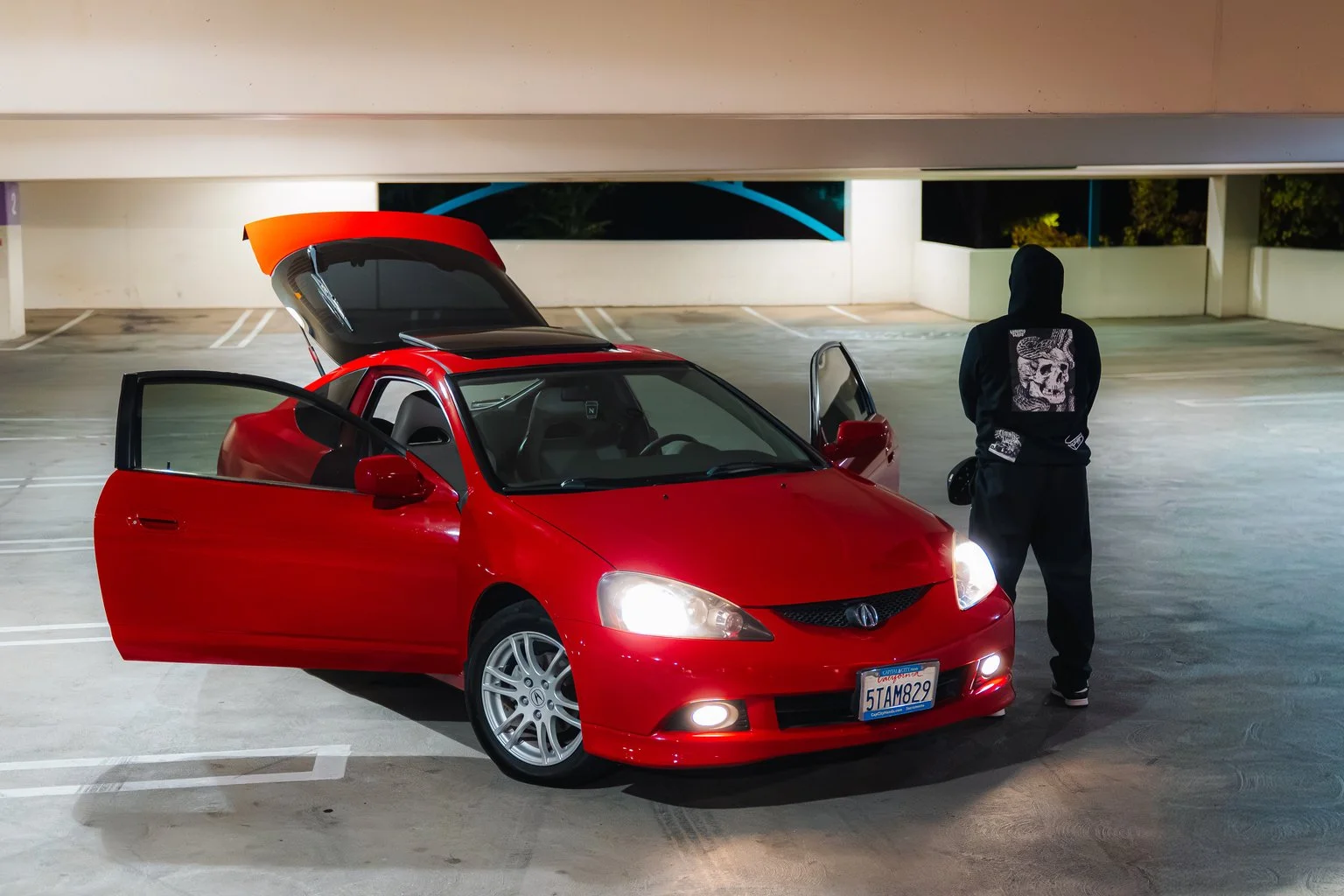 Red car with open front door and trunk parked in a mostly empty parking lot at night. A person wearing dark clothing and a hoodie stands next to the car.
