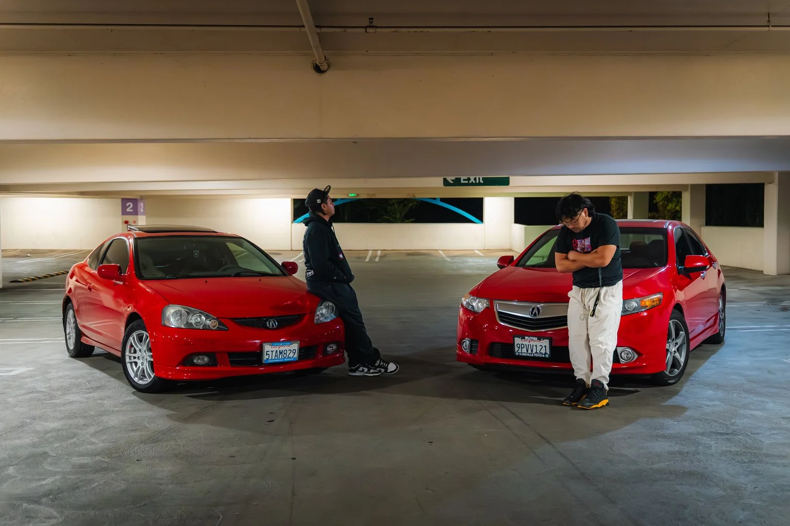 Two young men with red cars in an underground parking lot at night, one leaning against the car and the other standing with arms crossed, both looking away.
