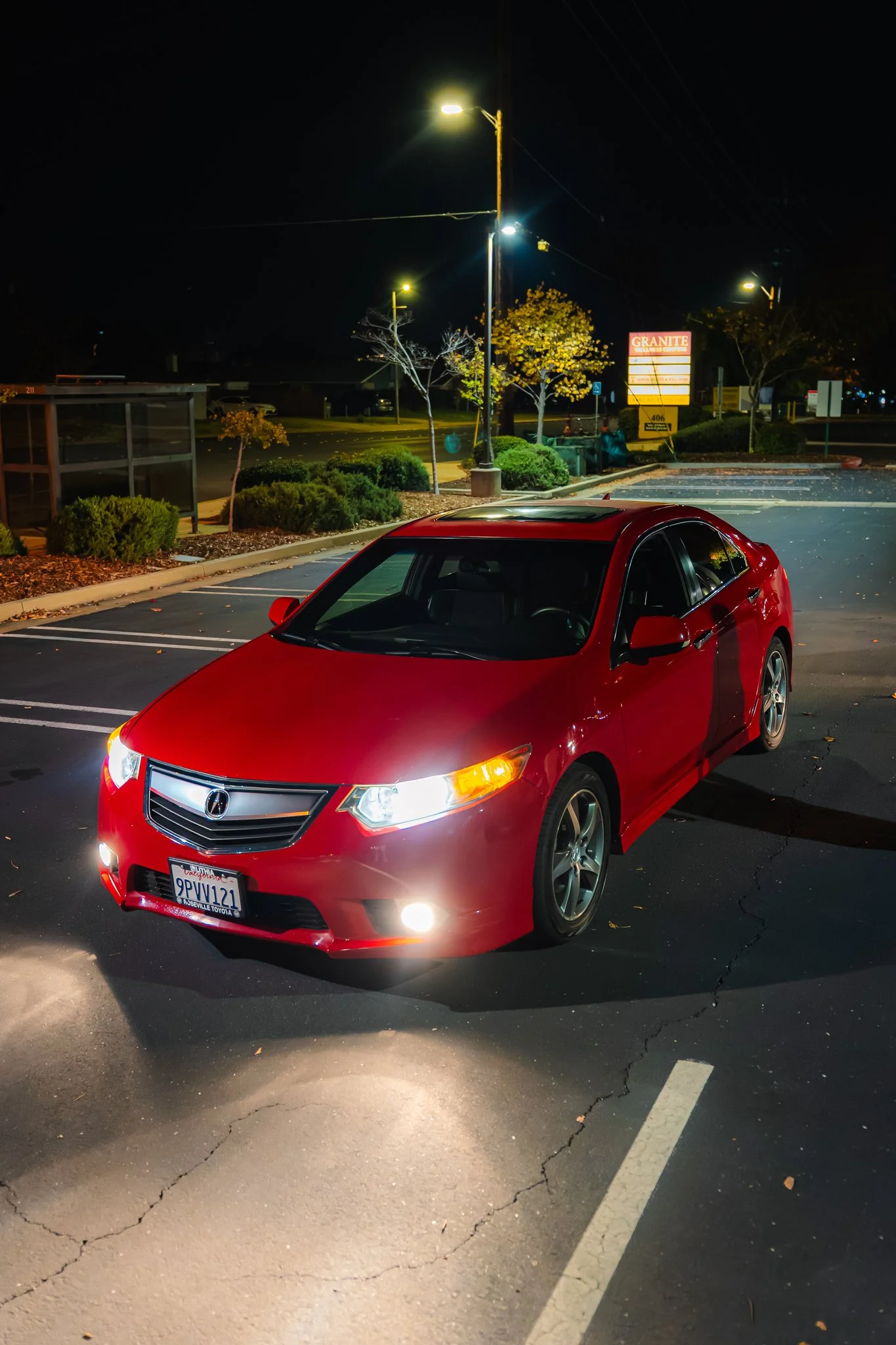 Red Acura car parked in a mostly empty parking lot at night, illuminated by streetlights, with trees and a business sign in the background.