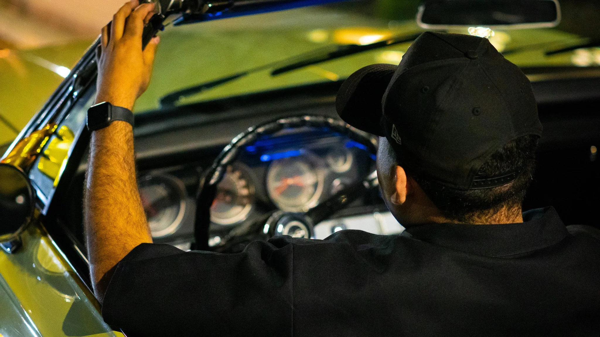 A man wearing a black cap and black shirt is driving a vehicle. His right hand is on the steering wheel, and he is looking forward. The dashboard and dashboard gauges are visible inside the car.