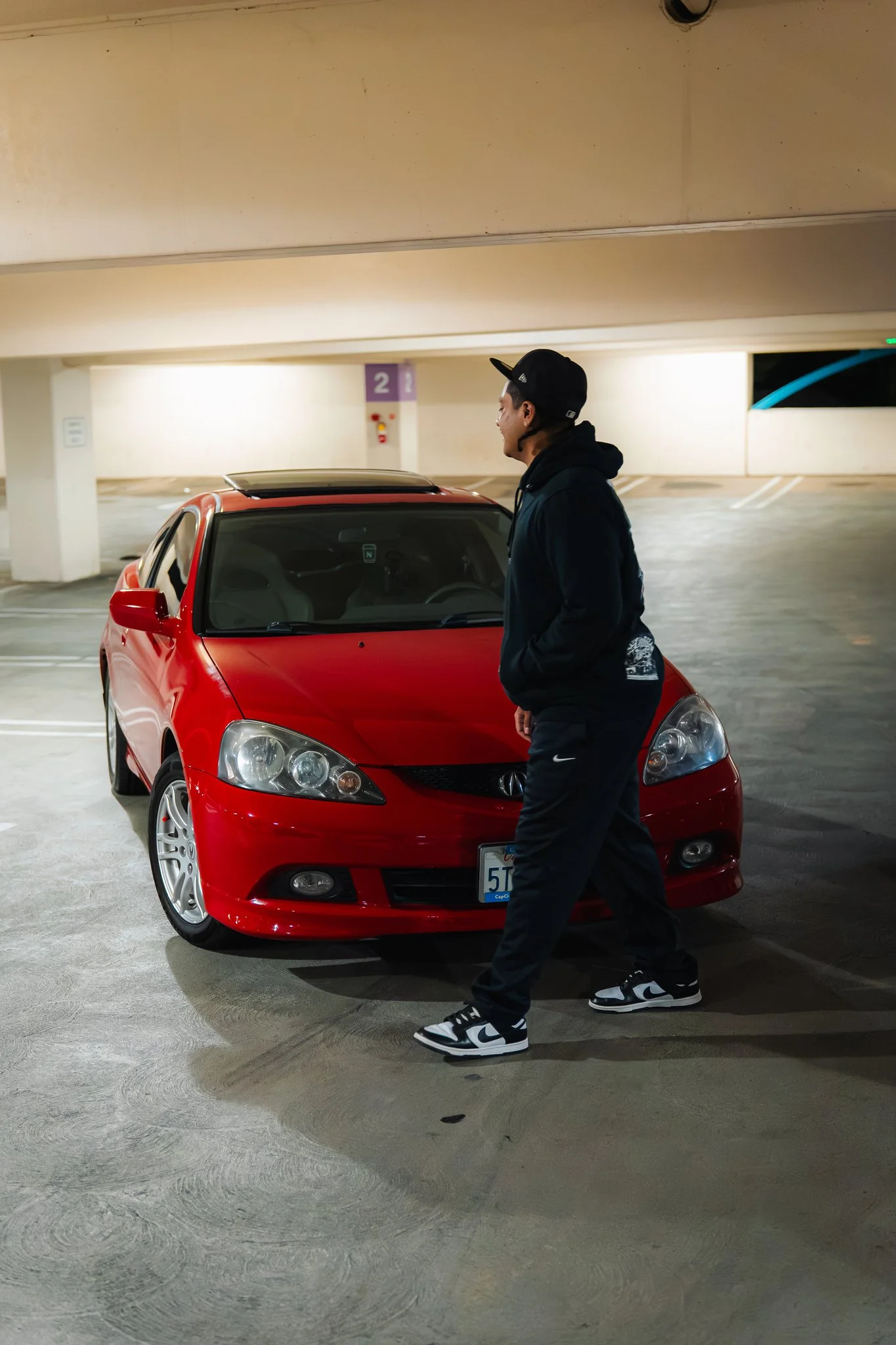 A man wearing a black hoodie, black pants, and black-and-white sneakers walking past a red car in a parking garage.