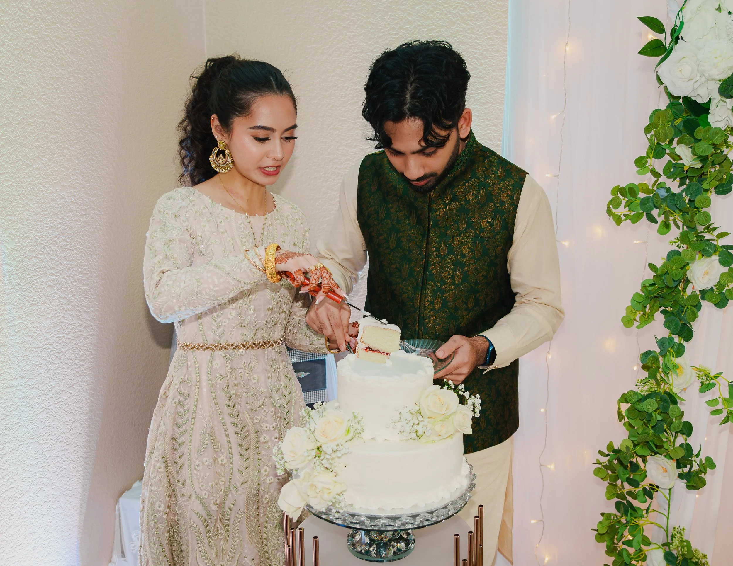 A couple dressed in traditional Indian attire cutting a wedding cake at a celebration.