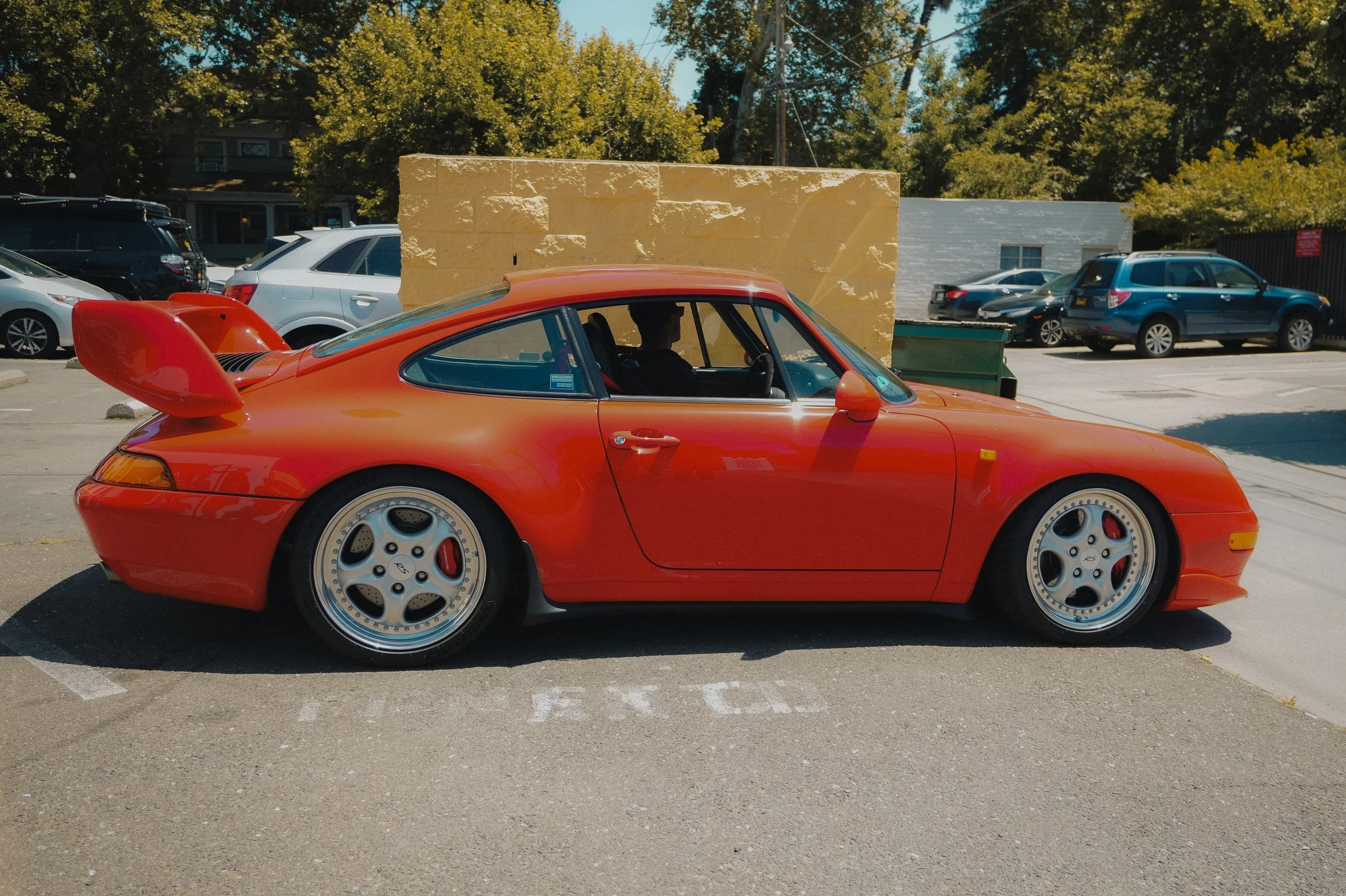 A red sports car parked in a parking lot with other cars, a brick wall behind it, and trees and buildings in the background.