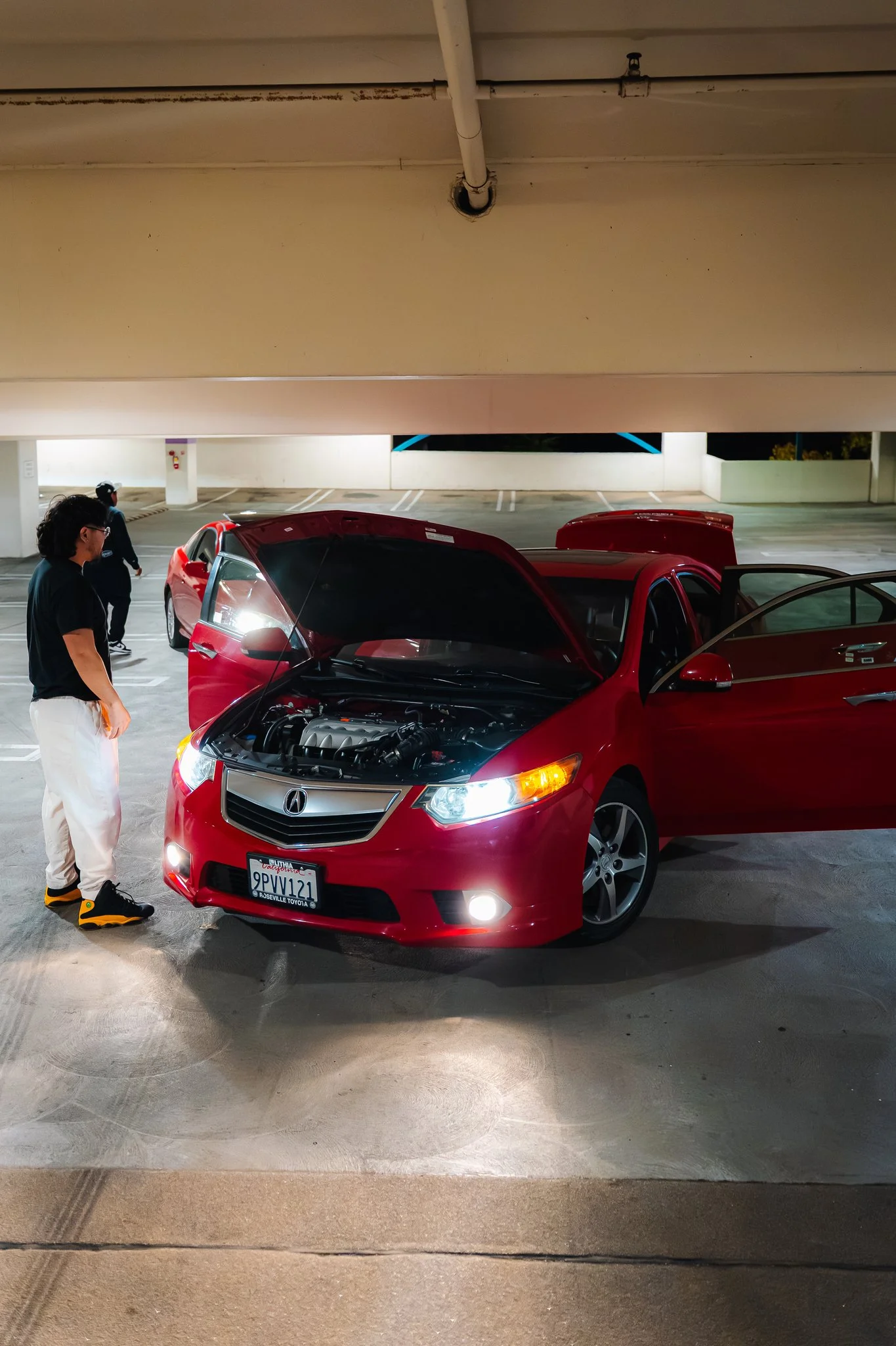 A red car with an open hood parked in a parking garage at night, with two men standing beside it and looking inside.