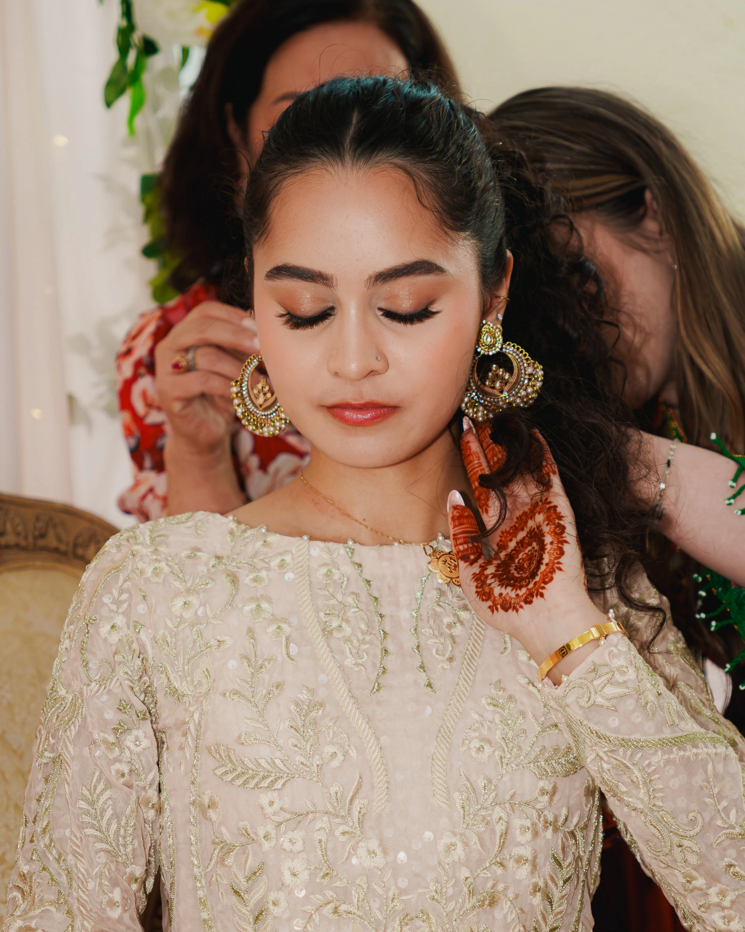 A woman with closed eyes wearing traditional jewelry and a cream-colored embroidered dress, while another woman in background adjusts her earring.