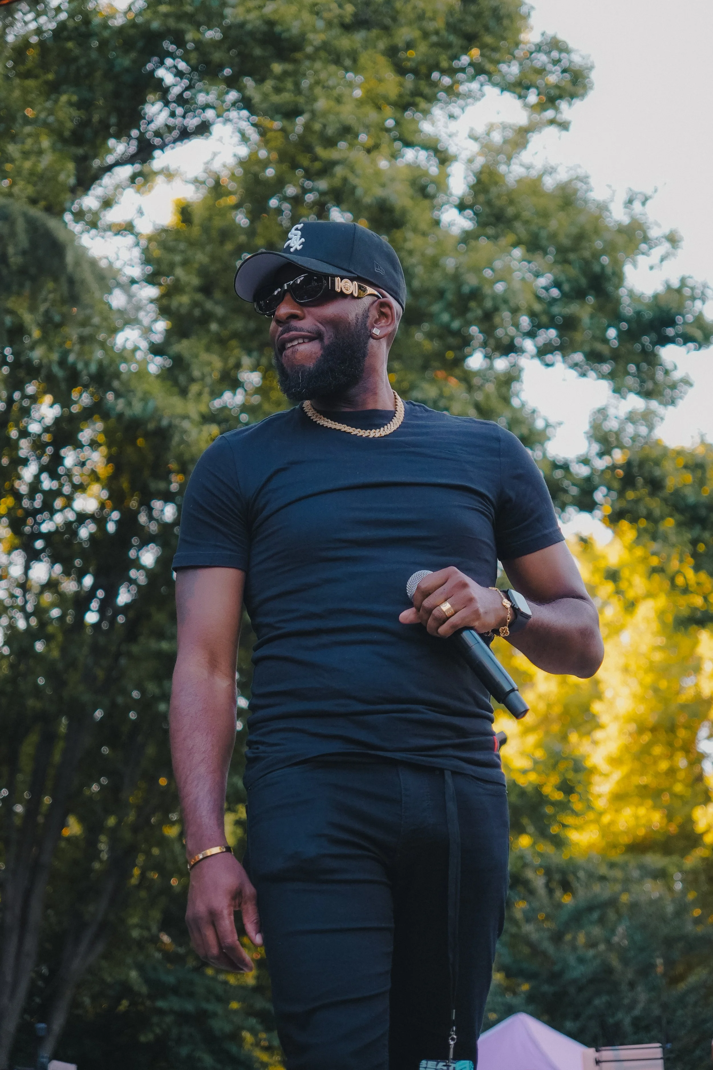 Man with beard wearing black t-shirt, black pants, black cap with white logo, sunglasses, gold chain, and earrings, holding a microphone outdoors in a park with trees in the background.