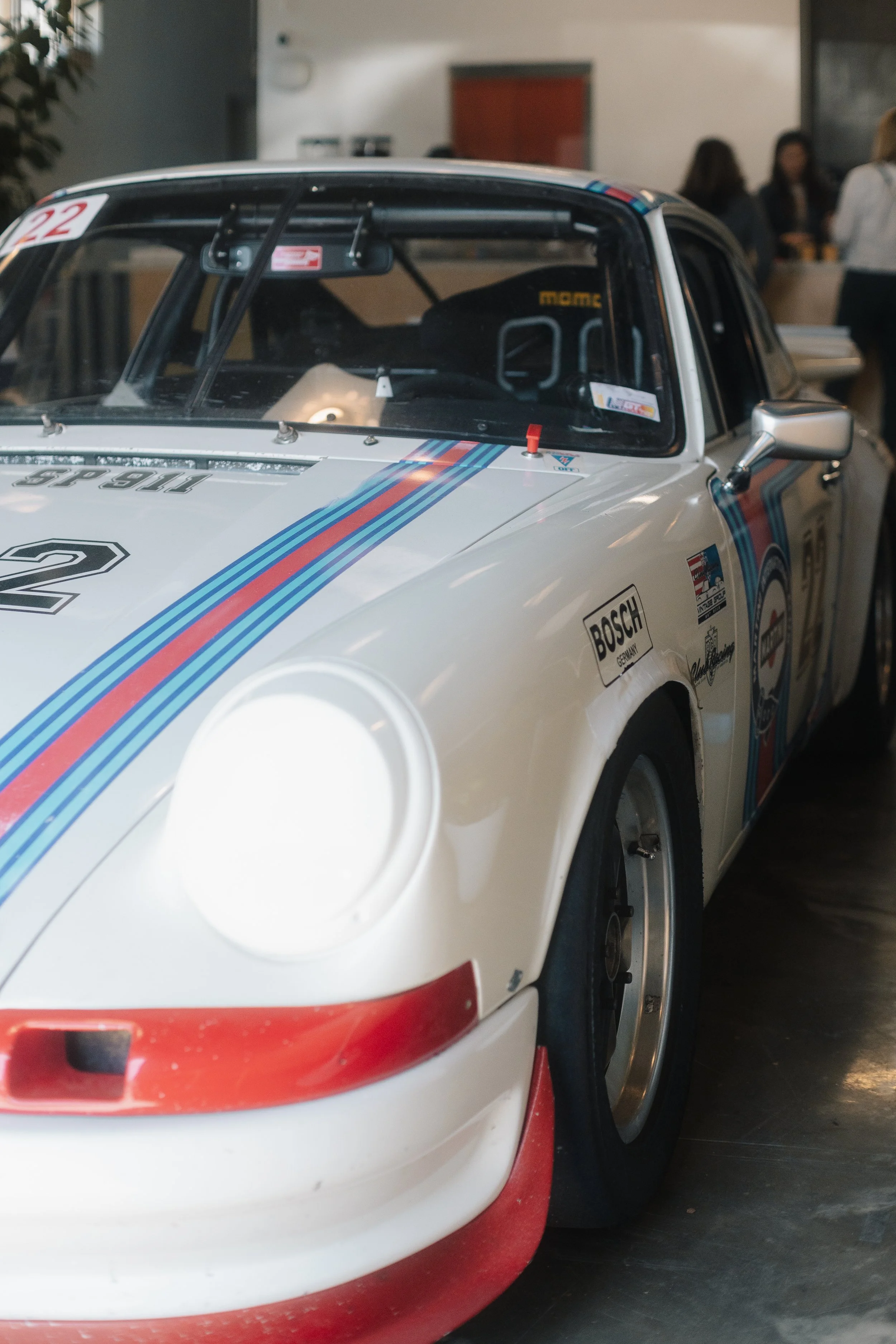 Close-up of a vintage white race car with red and blue stripes, number 2 on the hood, and sponsorship stickers, parked indoors with a group of people in the background.