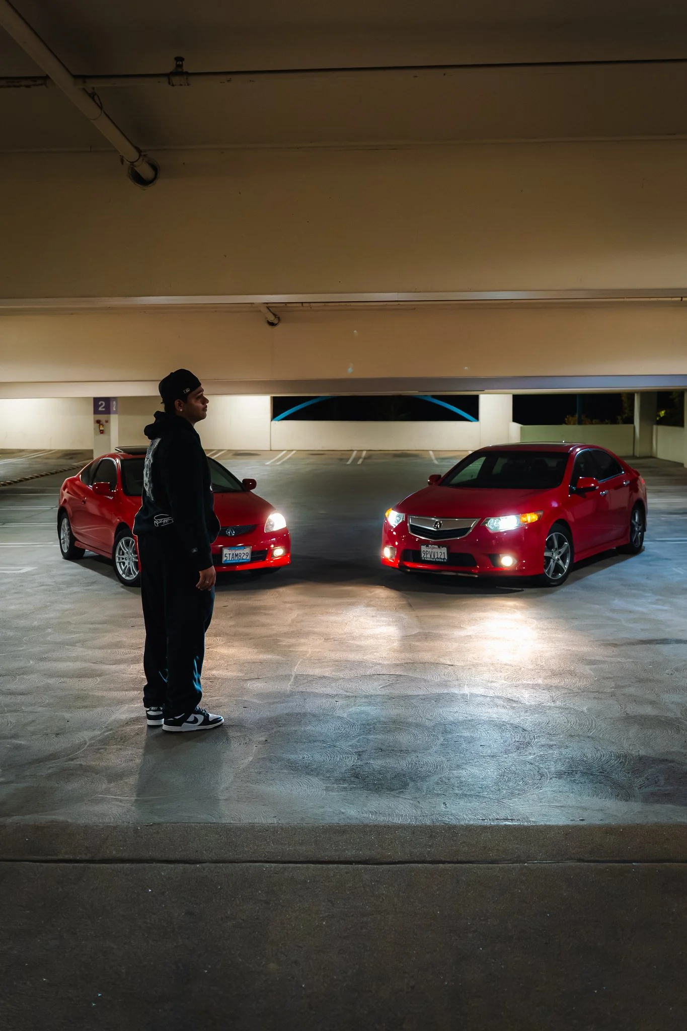 A young man standing in an empty parking garage at night, with two red cars parked behind him and the ceiling above.