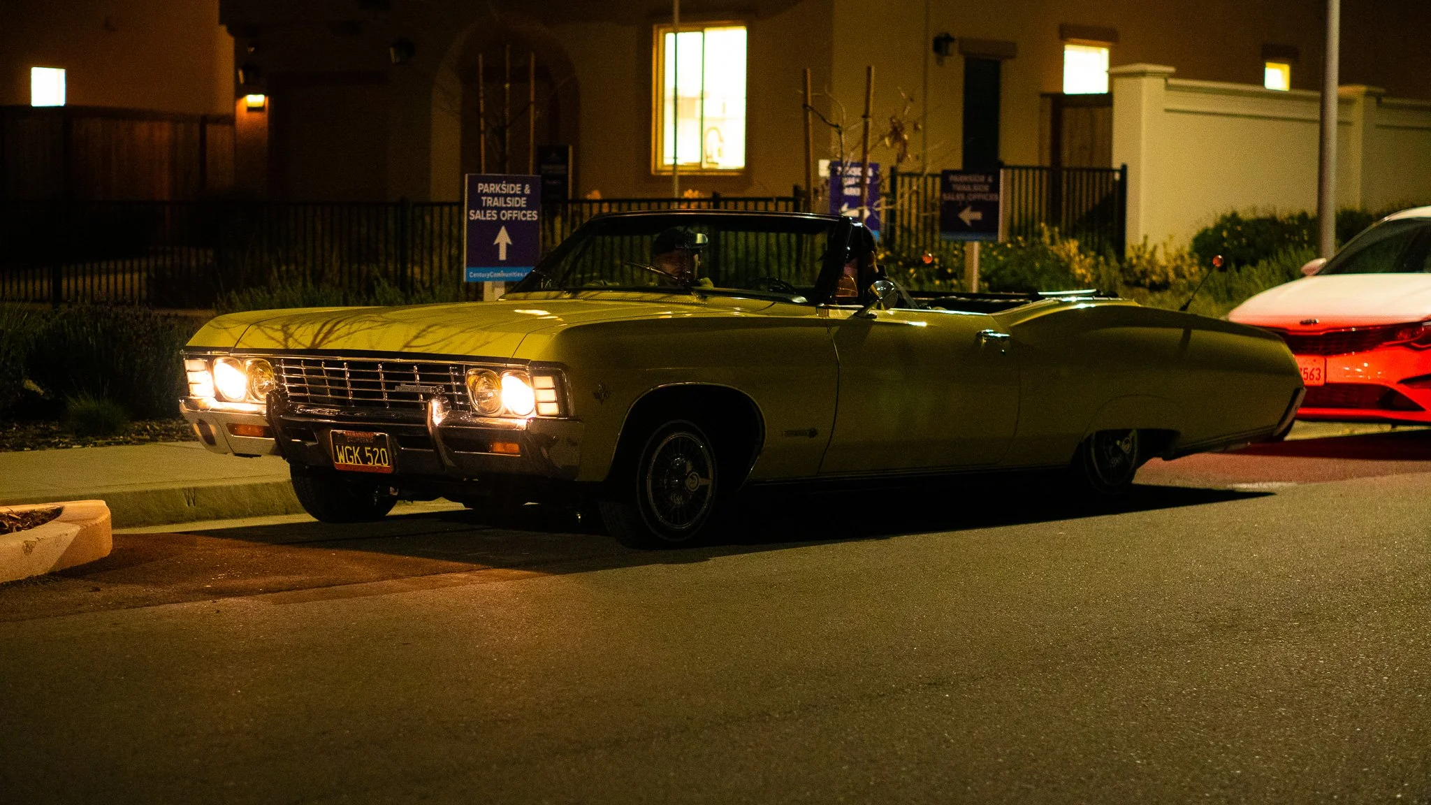 A vintage yellow convertible car parked on the street at night, with another modern white car partially visible behind it. There are signs in the background pointing to park and trailside sales offices, with illuminated windows and a fenced area.
