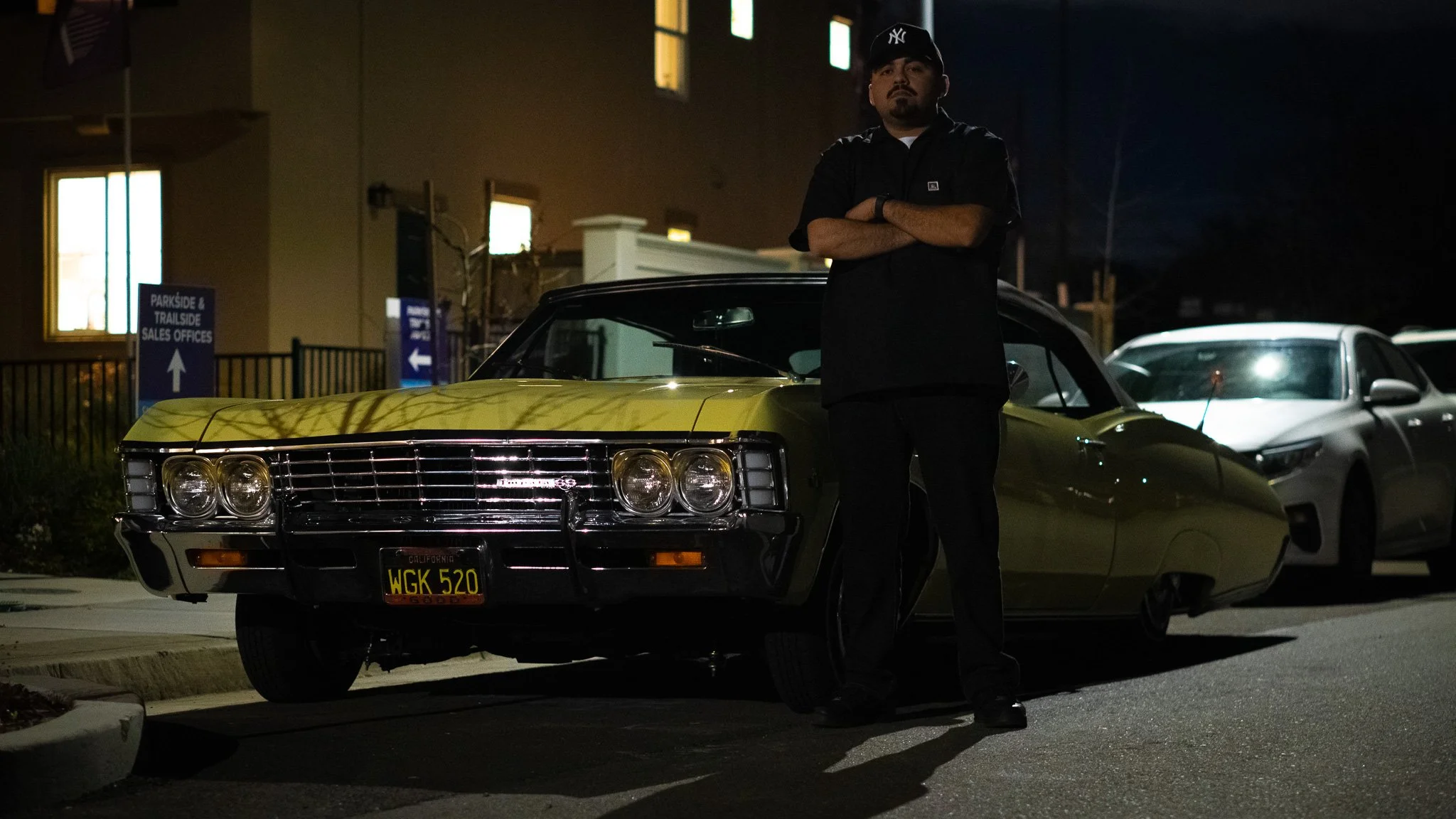 A man with crossed arms standing next to a green vintage Chevrolet Monte Carlo car at night. The man is wearing a black baseball cap and a black shirt, and there are other cars and buildings in the background.