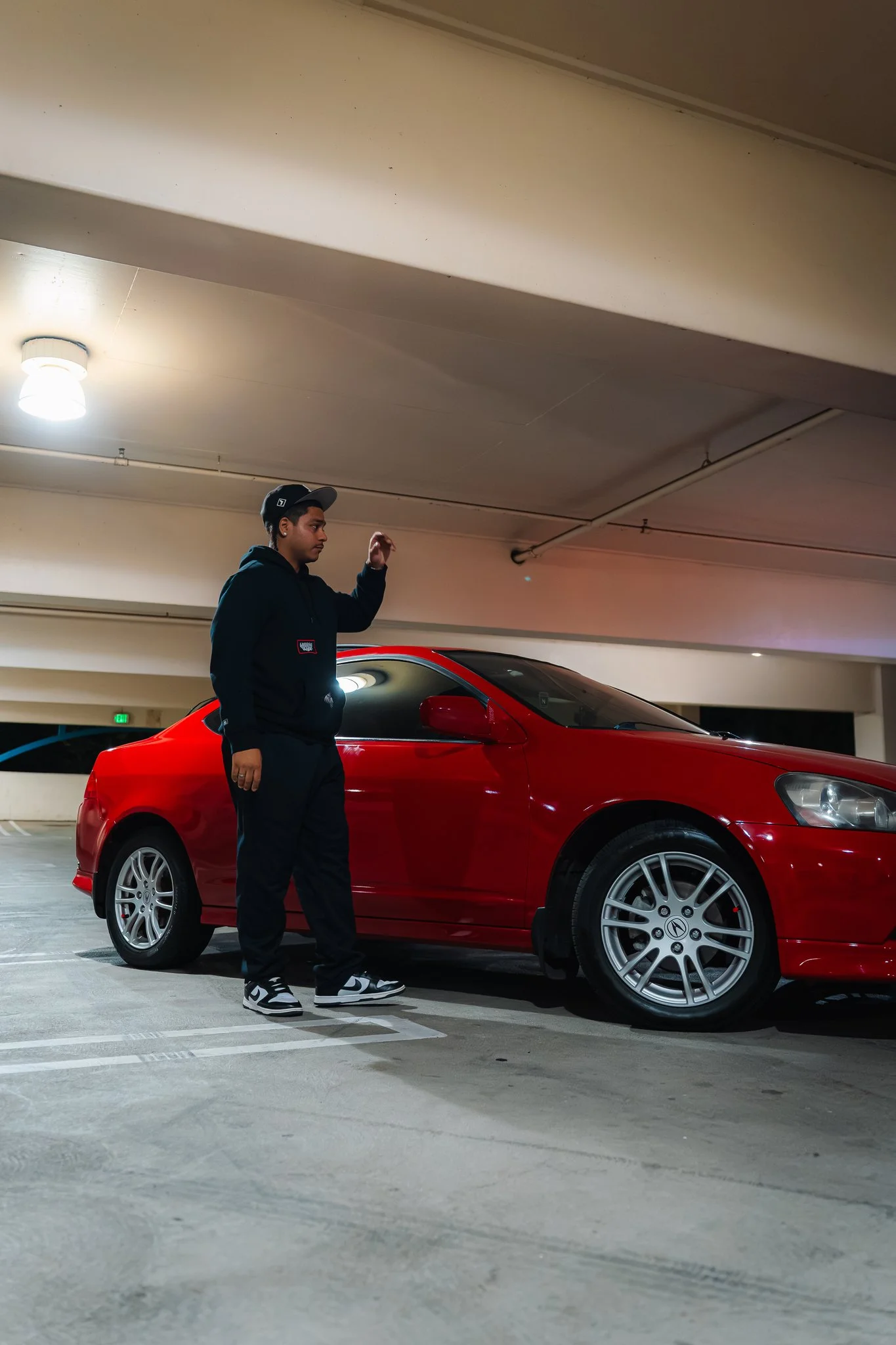 A young person wearing a black hoodie, black pants, and sneakers, standing beside a red car in an indoor parking garage.