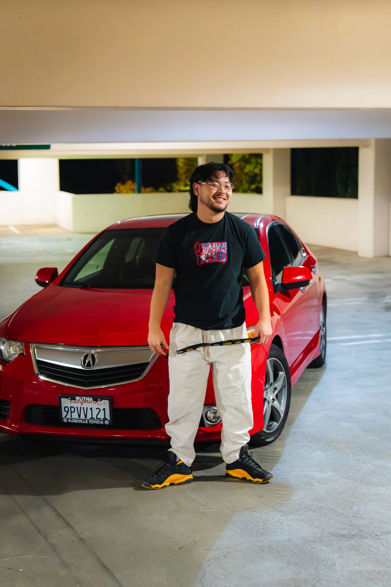 A man with glasses and a beard smiling, standing in front of a red car in a parking garage at night. He is wearing a black graphic T-shirt, beige pants, and black and yellow sneakers.