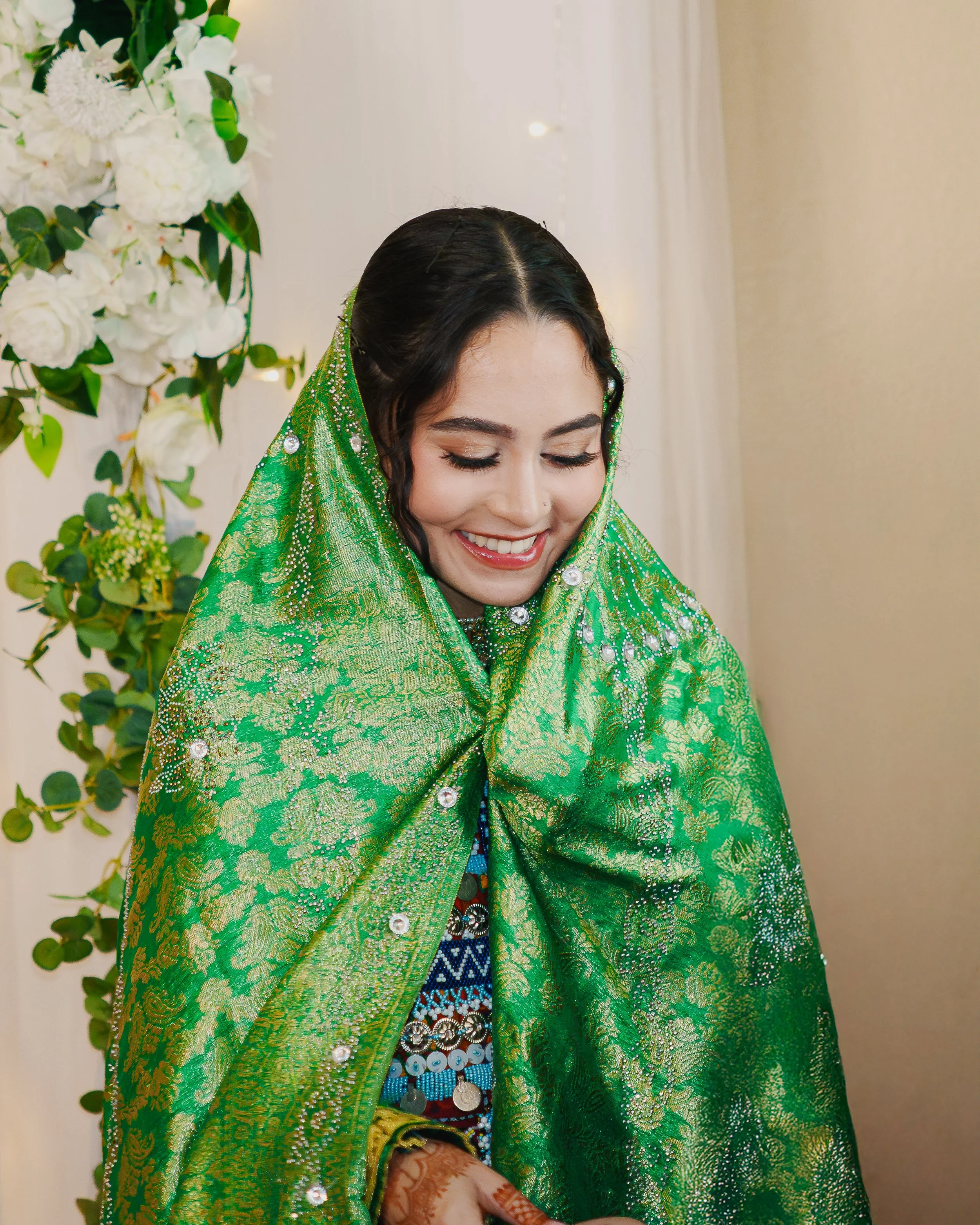 A woman dressed in a traditional Indian green saree, decorated with intricate gold patterns and embellished with mirrors and beads, is smiling with her eyes closed. She is wearing jewelry, including rings and a necklace, and has henna designs on her 