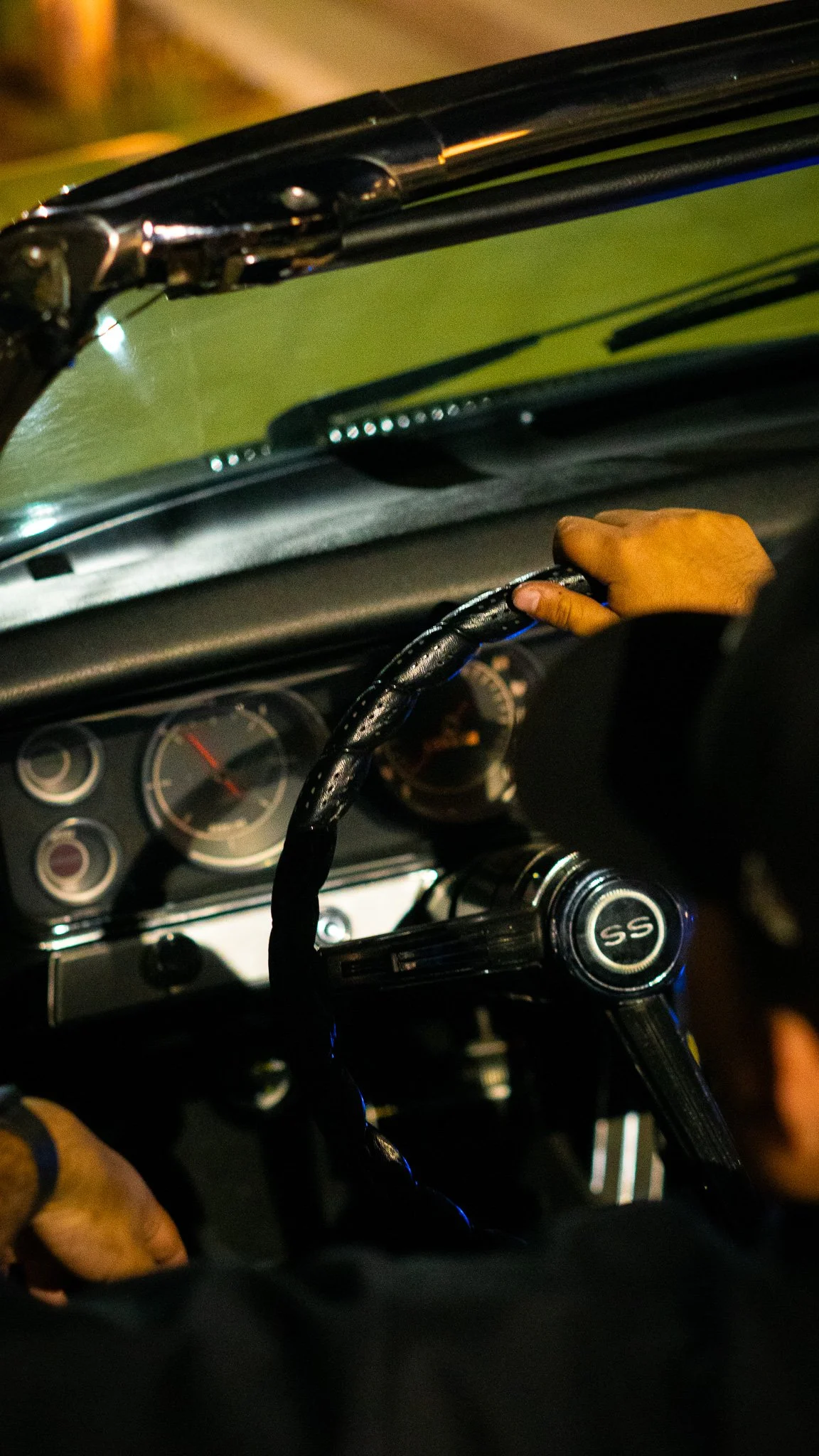 Inside a vintage car, showing the driver's hand on a steering wheel, dashboard, and windshield with a view of the road at night.