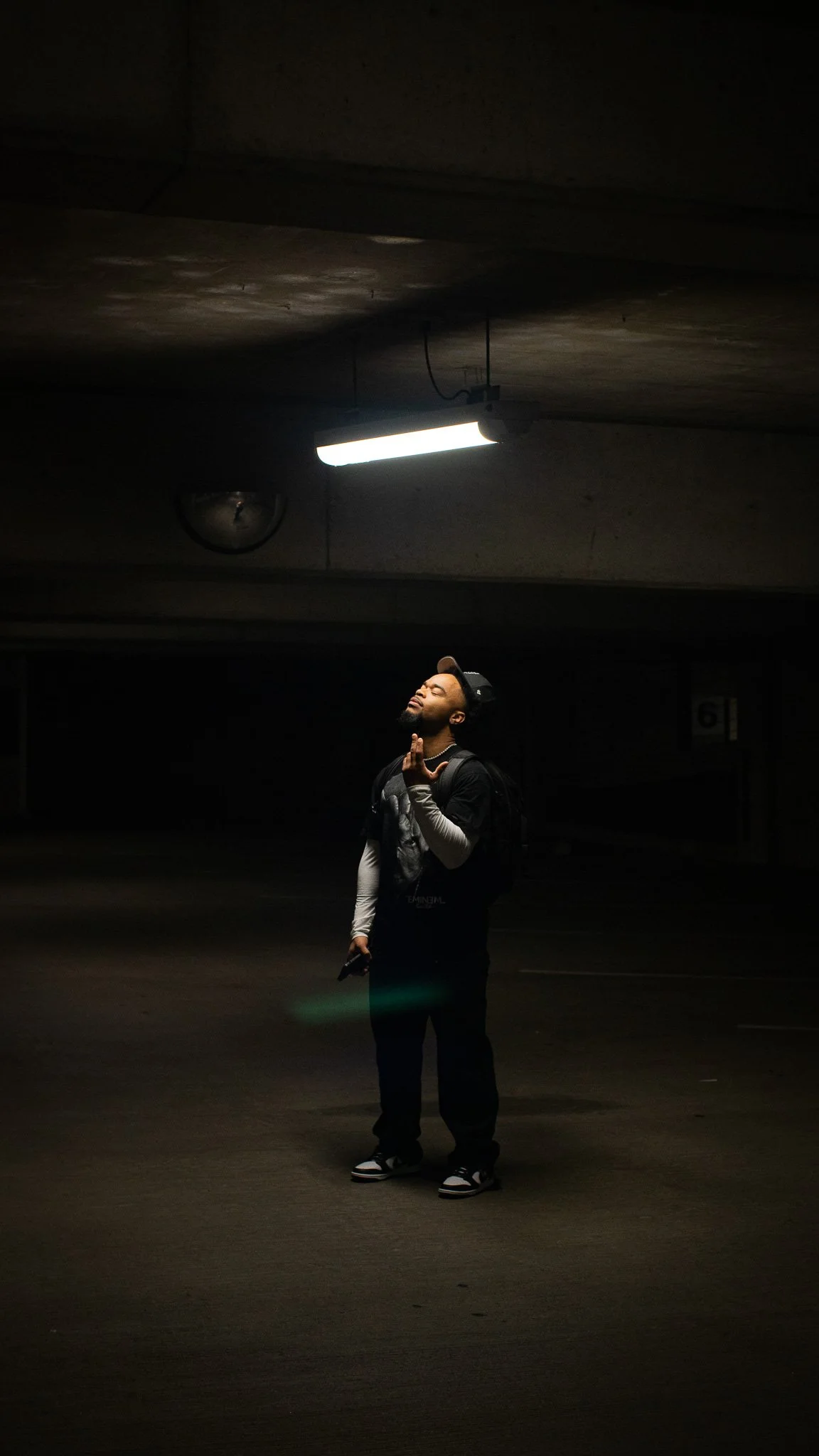 Young man with backpack and cap standing in a dimly lit parking garage, illuminated by a fluorescent ceiling light, with a contemplative expression.