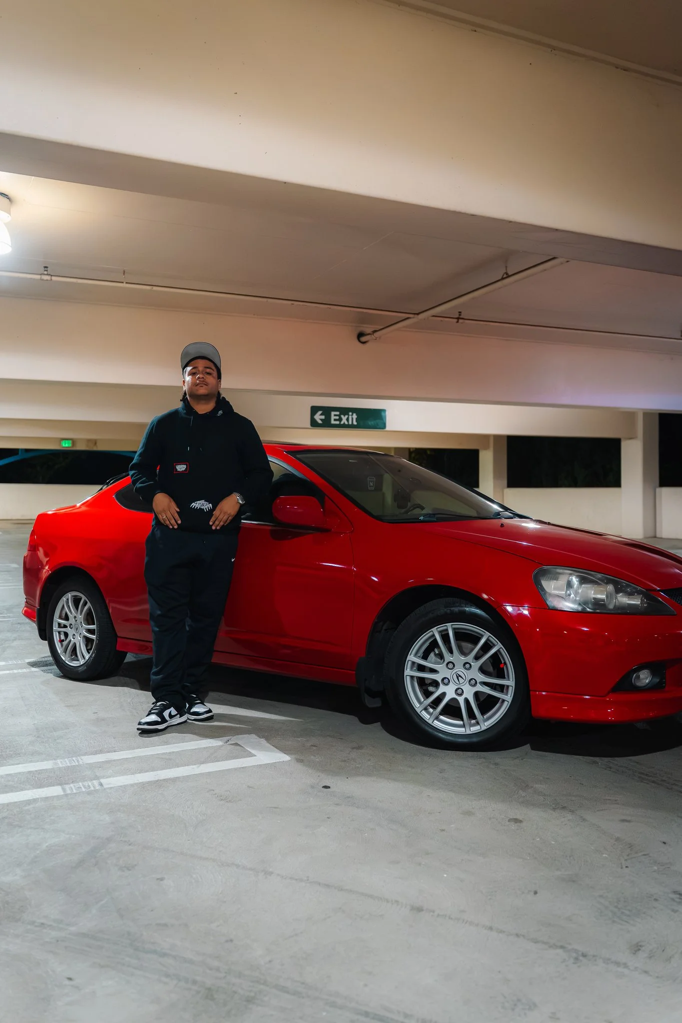 A young man wearing a black hoodie, black pants, and sneakers standing in front of a red car in an underground parking garage. There is a green exit sign on the wall behind him.