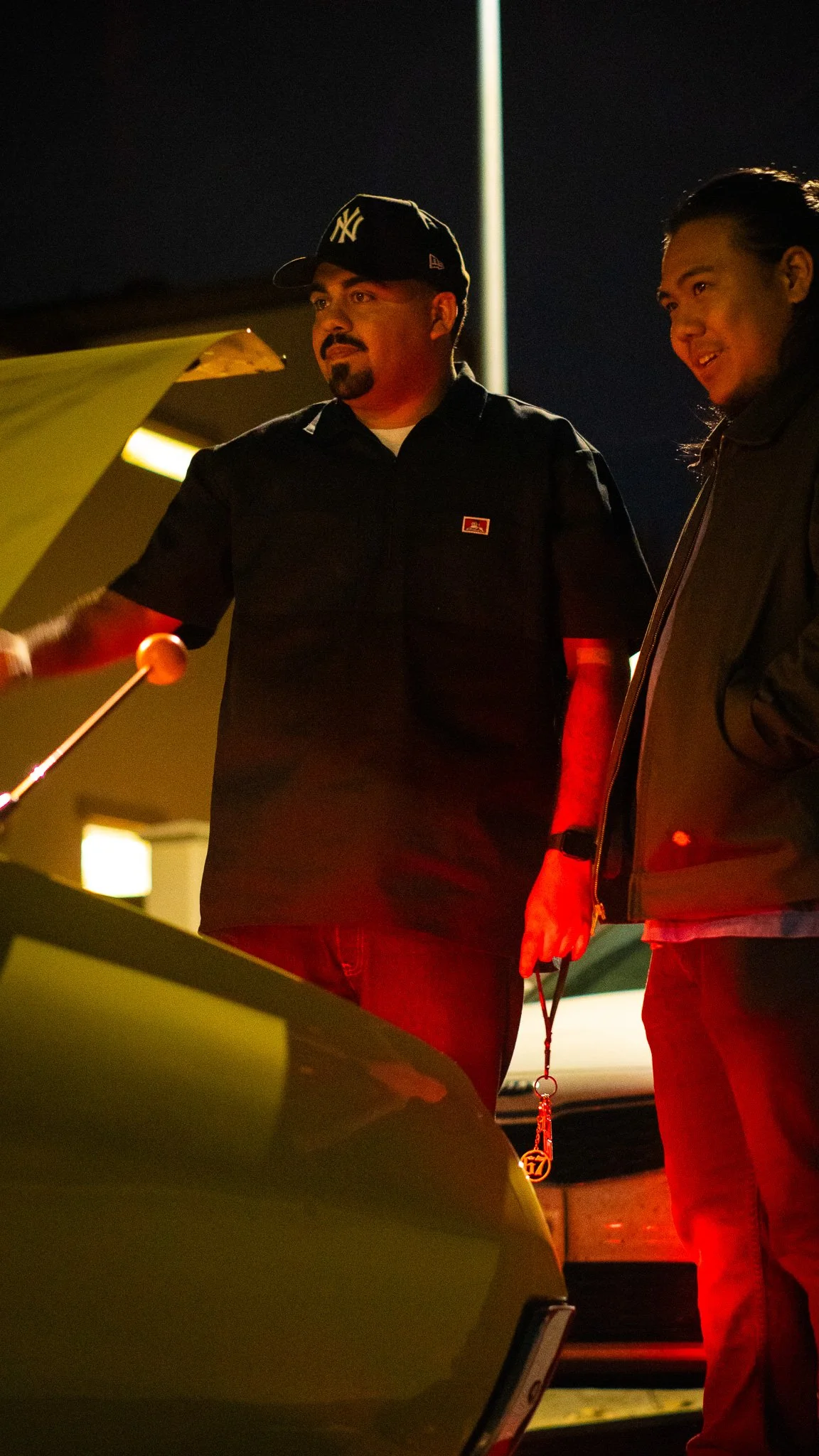 Two men interacting at night, one playing a green piano and the other standing nearby, holding a leash with a keychain, under a streetlight.