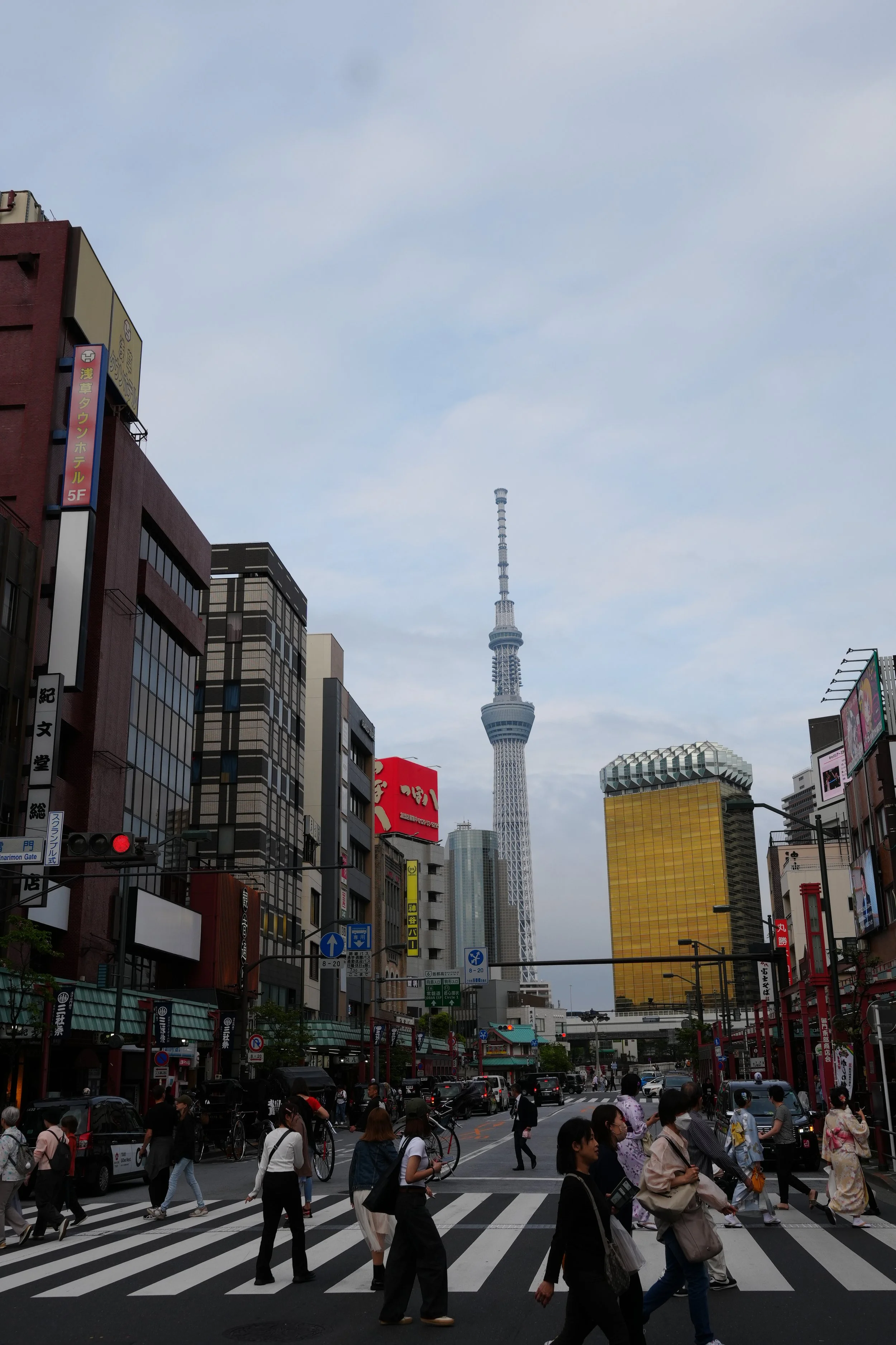 City street scene with pedestrians crossing at a crosswalk, tall buildings including Tokyo Skytree and other skyscrapers, and a cloudy sky overhead.