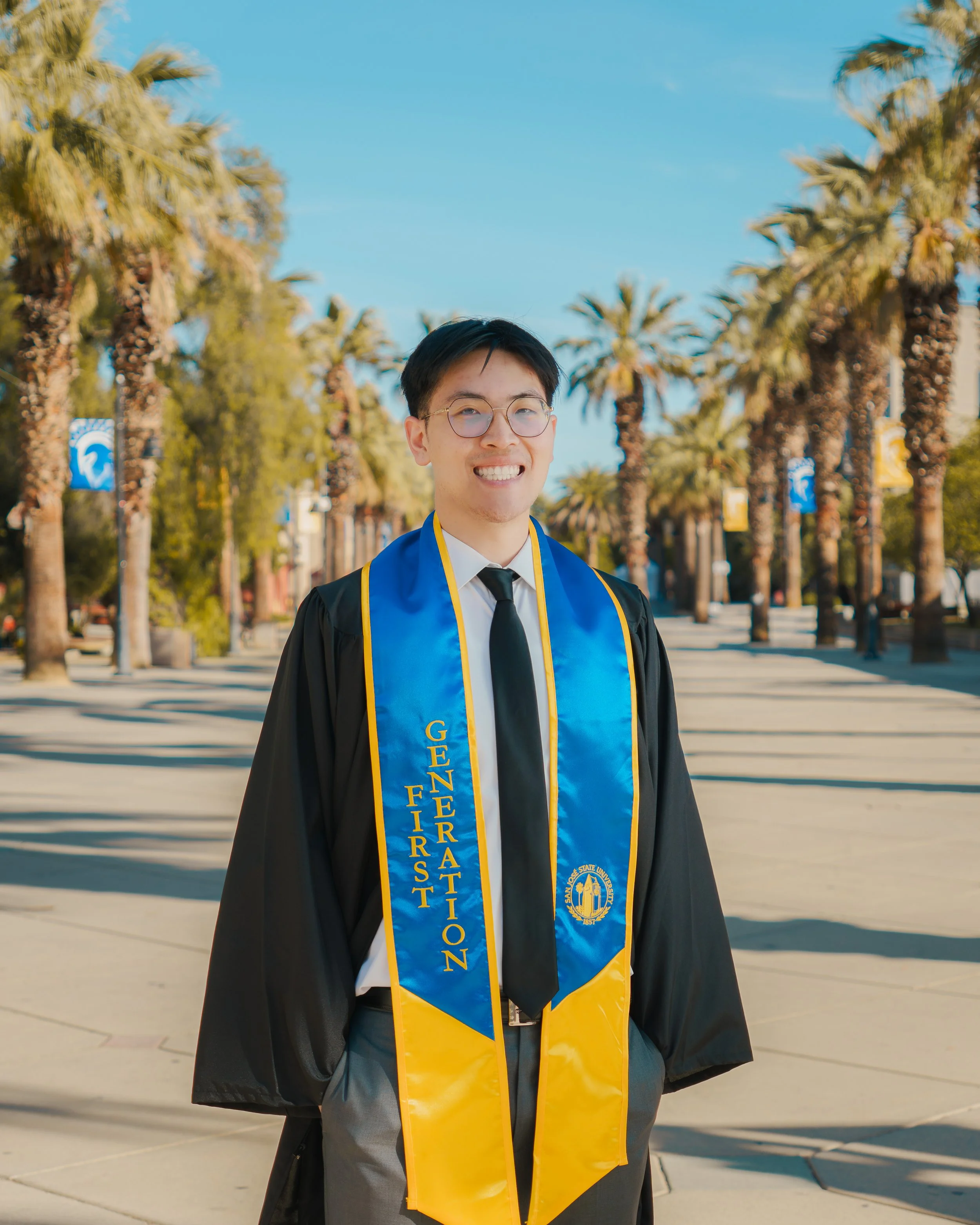 A young man wearing glasses in a graduation gown and stole with gold and blue colors, standing outdoors lined with palm trees and banners, smiling at the camera.