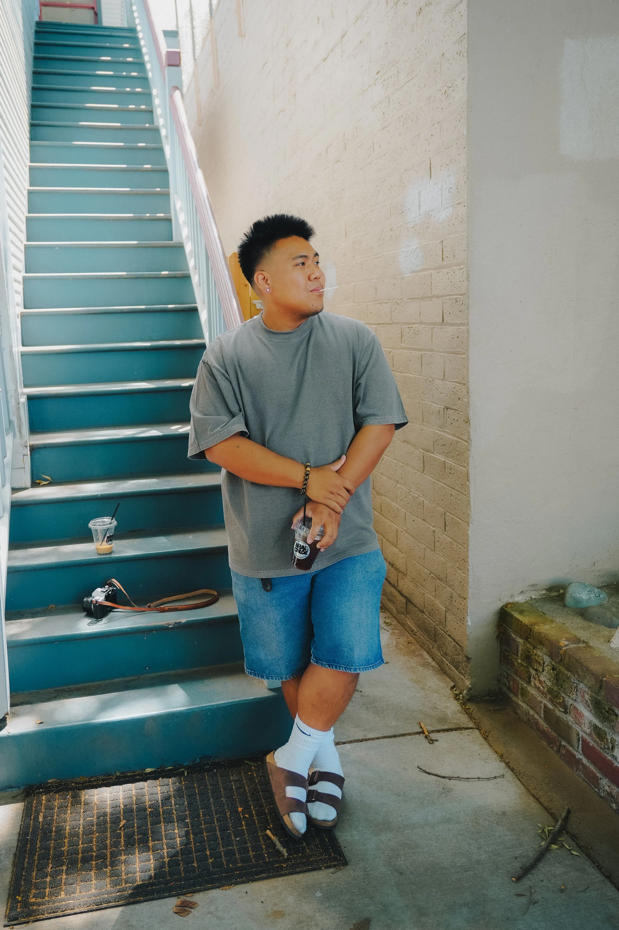 A young man standing on a sidewalk, leaning against a brick wall, holding an iced coffee, wearing a gray t-shirt, blue shorts, white socks, and black slides with white stripes, with a spiral bracelet on his right wrist, near a staircase with light bl