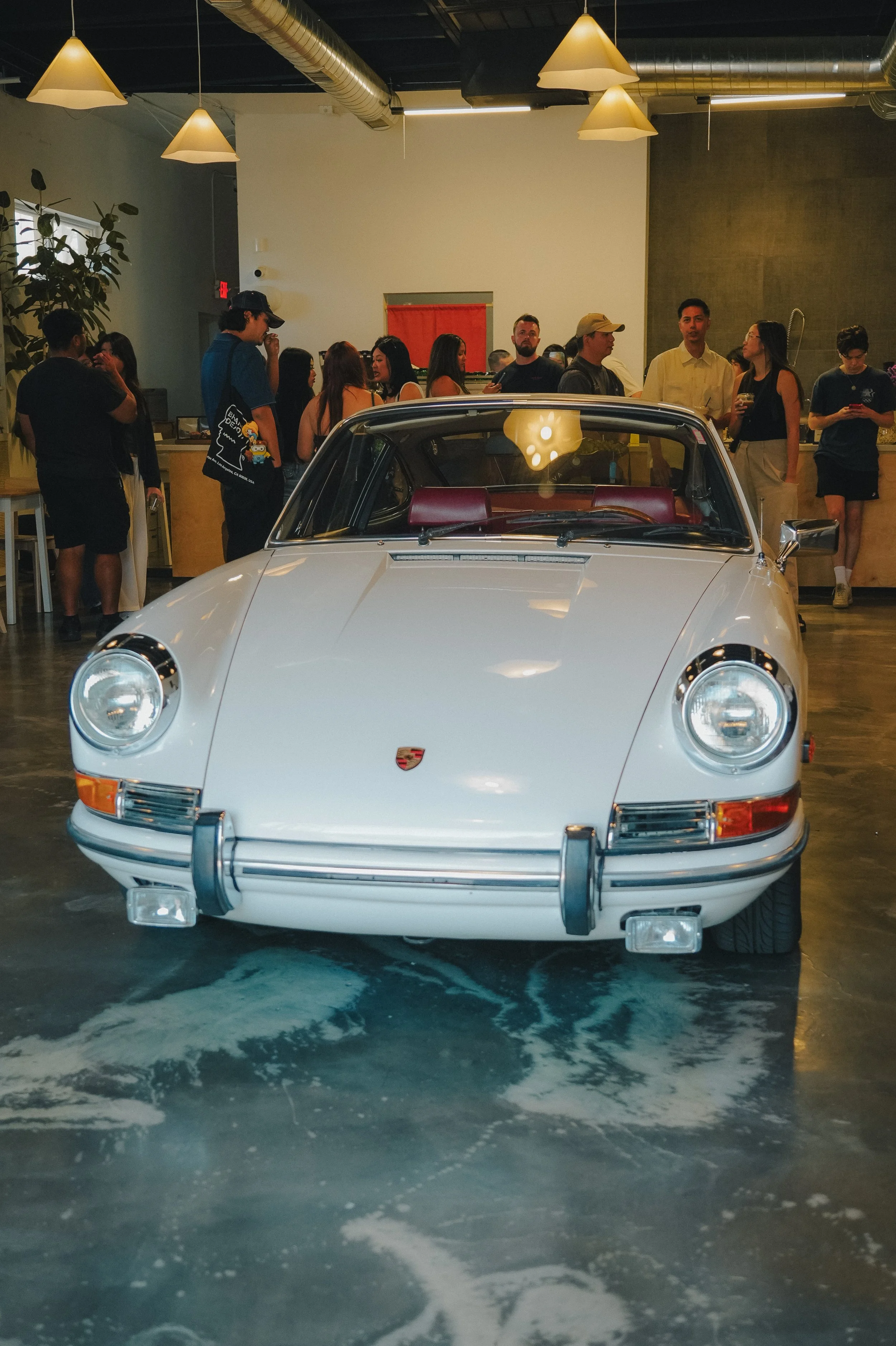 Front view of a white Porsche 911 sports car inside a modern indoor space, with a group of people in the background.
