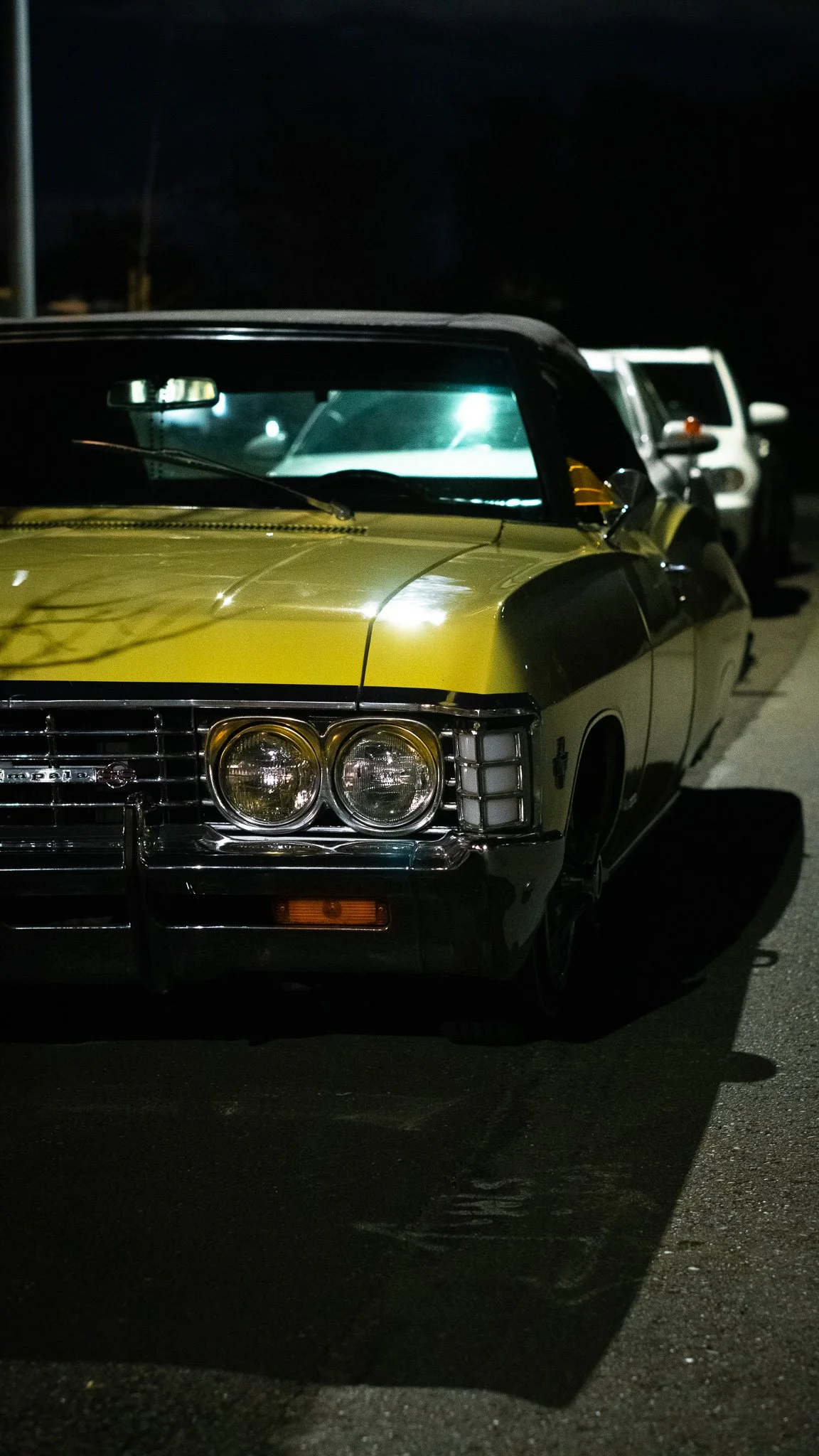 Nighttime photo of a vintage yellow and black classic car parked on the street, with two other vehicles visible behind it, all illuminated by streetlights.