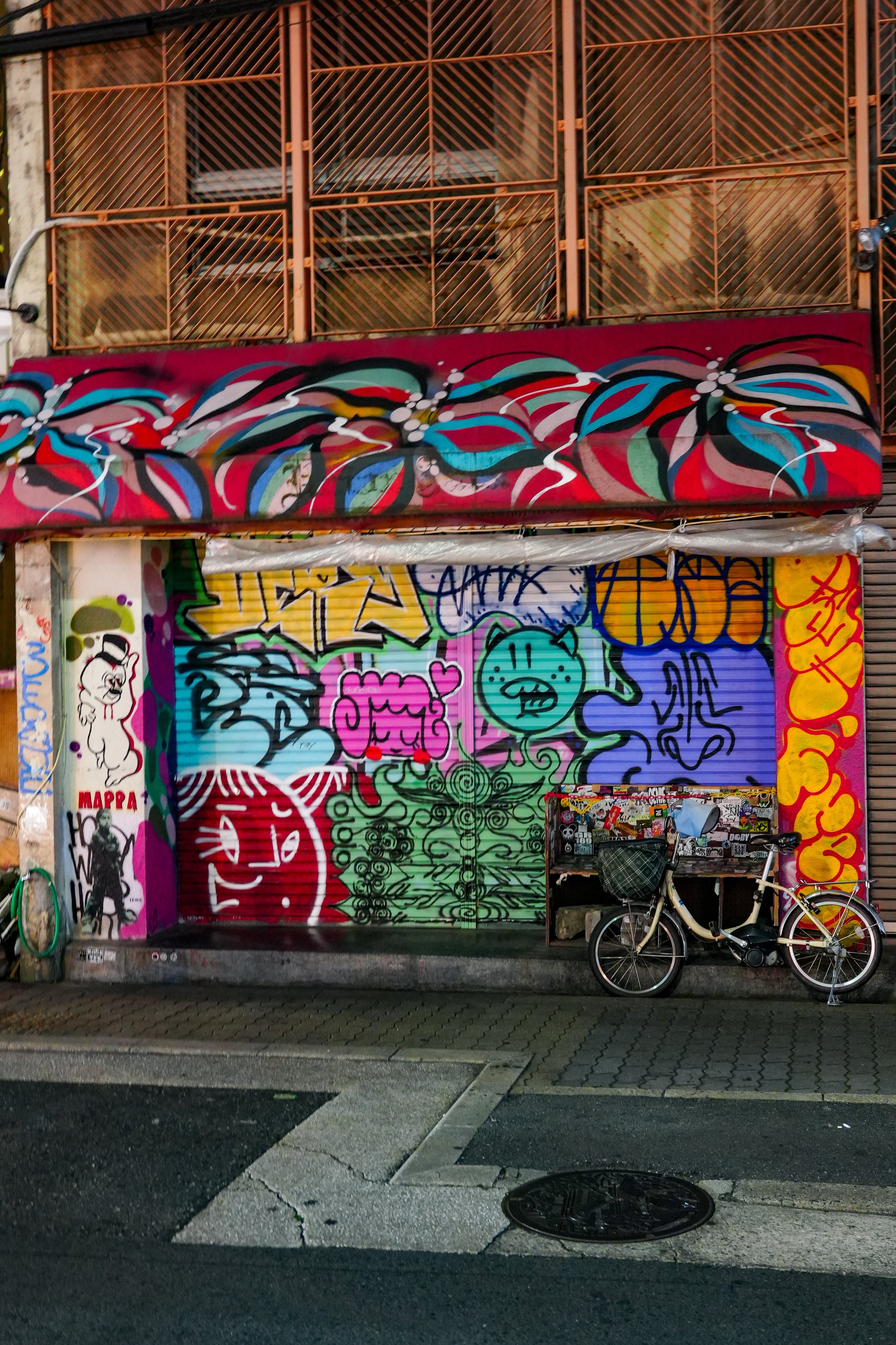 A storefront covered with colorful graffiti art including various characters and abstract designs. A bicycle is parked in front of the shop, which is closed with a rolled-down shutter.